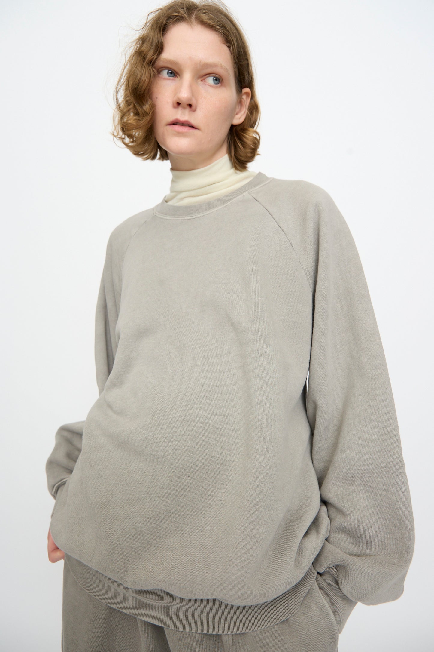 A person with short, wavy hair wears the Black Crane Raglan Sweatshirt in Stone—a loose-fitting gray cotton cashmere blend—over a beige turtleneck, paired with matching pants, and stands against a plain white background.