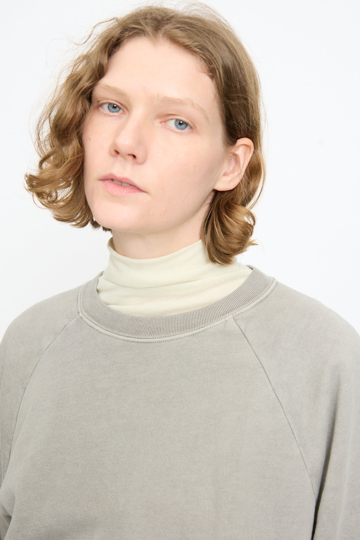 A person with short, wavy light brown hair is wearing the Black Crane Raglan Sweatshirt in Stone—crafted from organic cotton—layered over a white turtleneck, standing against a plain white background.