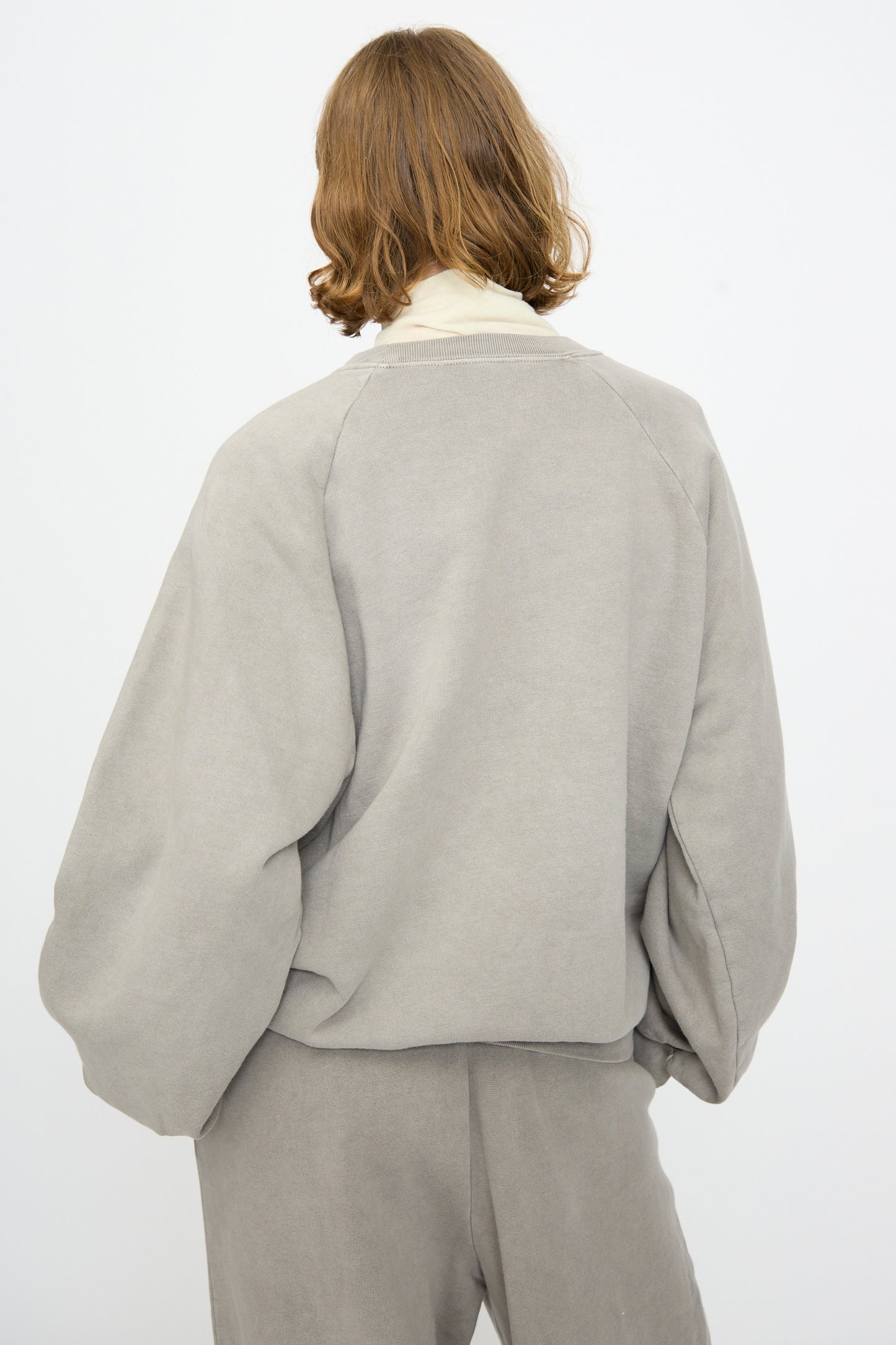 A person with shoulder-length light brown hair is shown from the back against a plain white background, wearing the Black Crane Raglan Sweatshirt in Stone—a loose, organic cotton grey pullover—paired with matching pants.