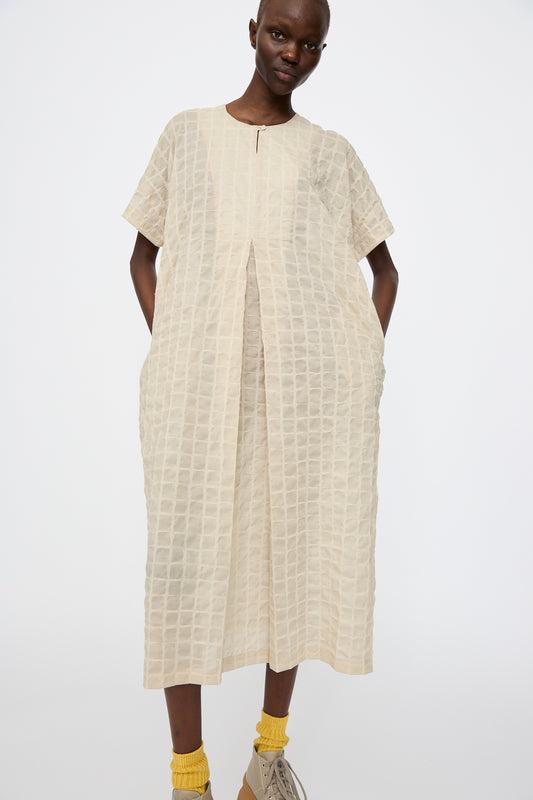 A person wears the Black Crane Ripple Finish Crew Neck Dress in Beige with pockets, paired with yellow socks and beige shoes, standing against a plain white background.