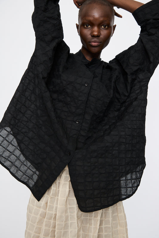 A person wearing the Black Crane Ripple Finish Shirt in Black, a loose-fitting Japanese cotton top with wide sleeves and a band collar, paired with a cream skirt, stands with arms raised against a plain background.
