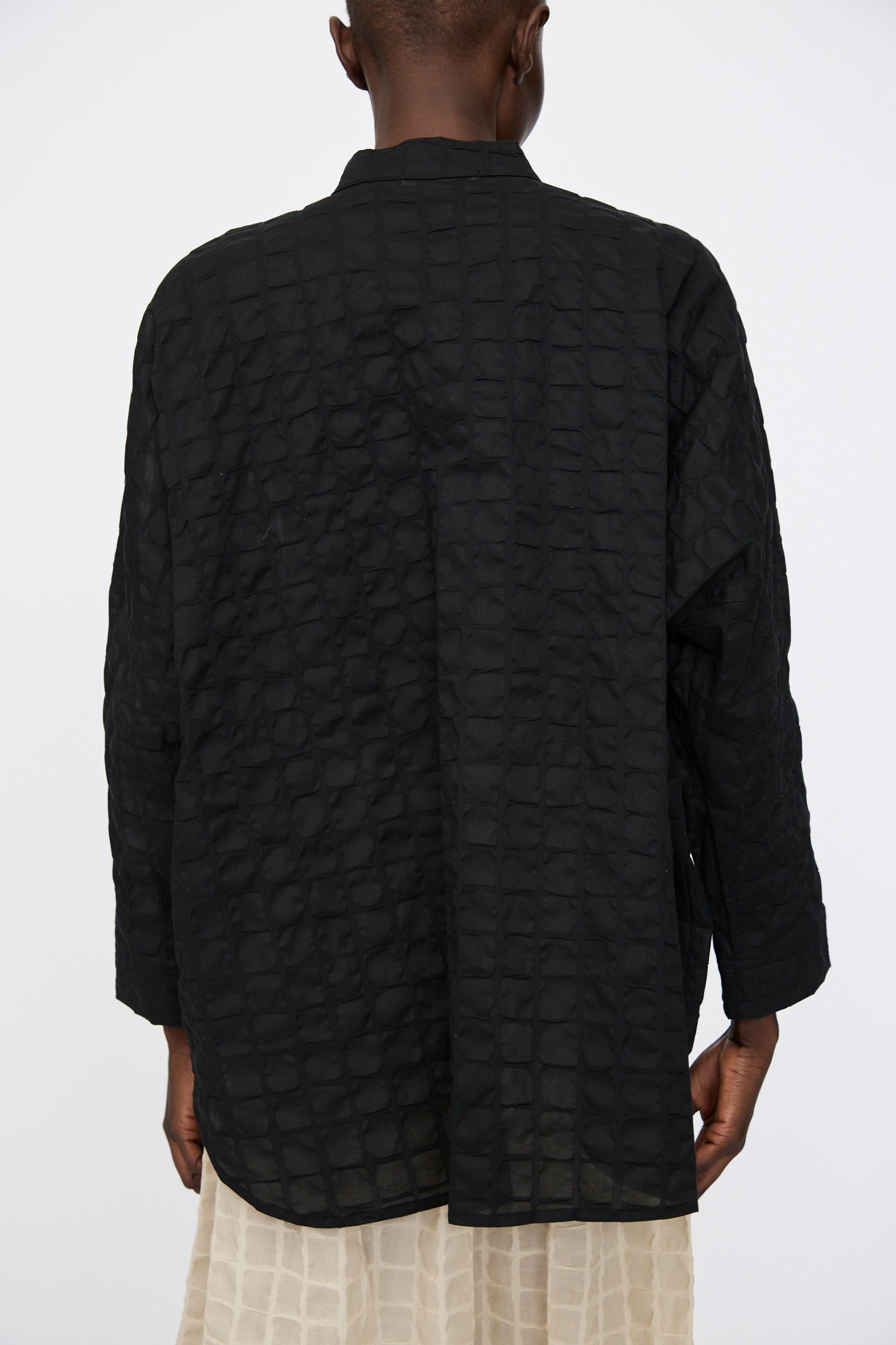 A person stands facing away, wearing the Black Crane Ripple Finish Shirt in Black with long sleeves and a band collar, paired with a cream skirt against a plain background.