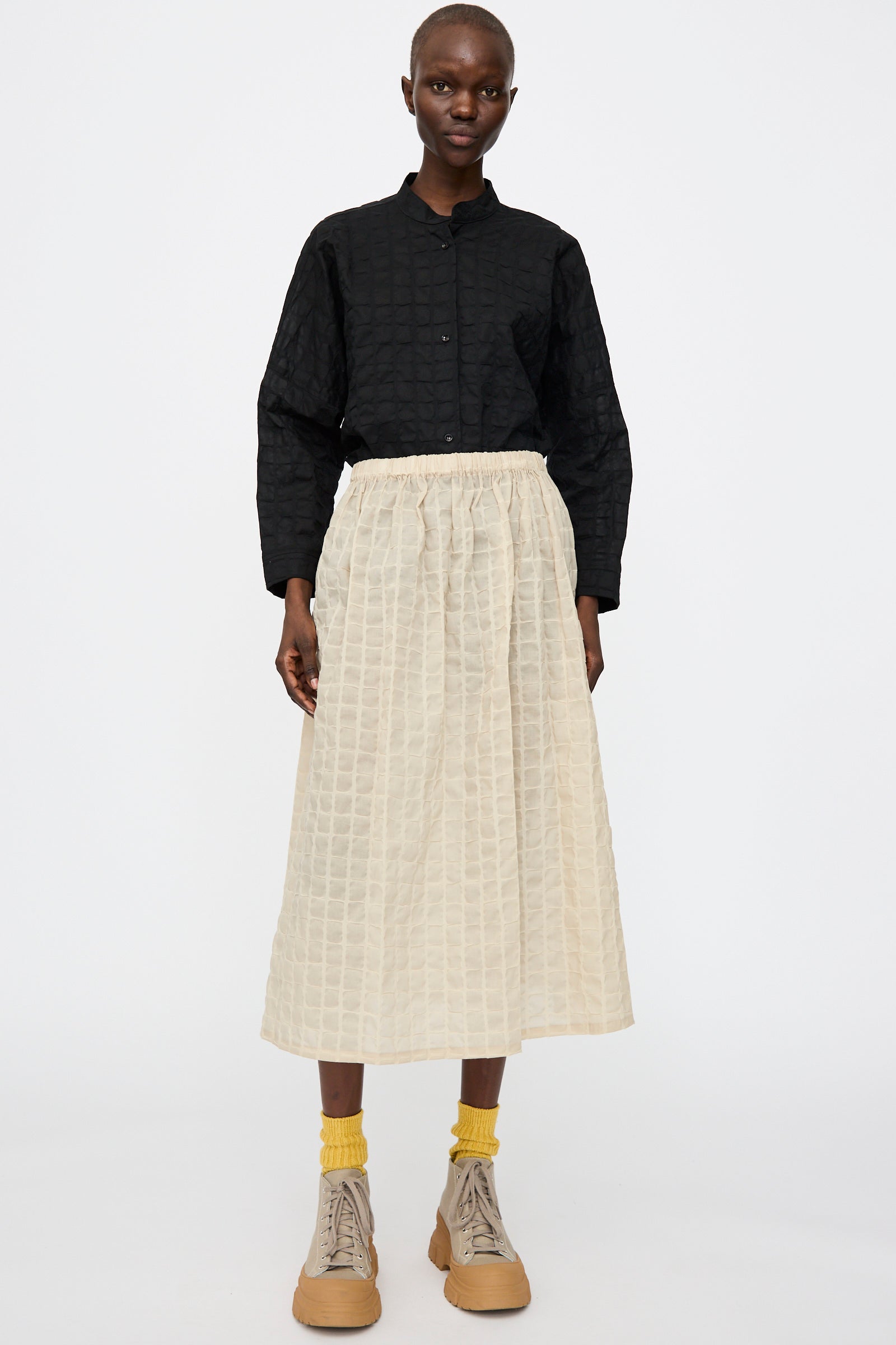 A person stands against a plain background wearing a textured black long-sleeve shirt, Black Crane's Ripple Finish Skirt in Beige, yellow socks, and beige lace-up shoes.