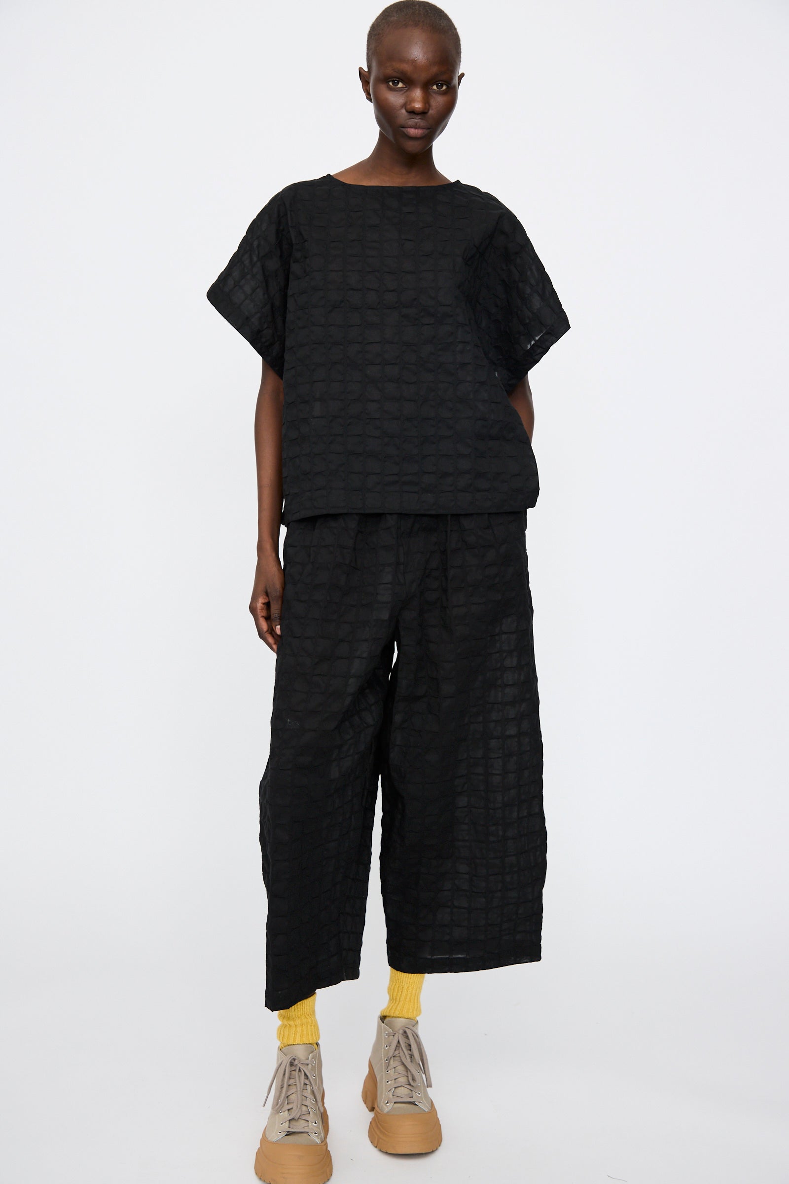 A person stands against a plain background, wearing Black Crane's Ripple Finish Top in Black with matching wide-leg pants, yellow socks, and beige sneakers.