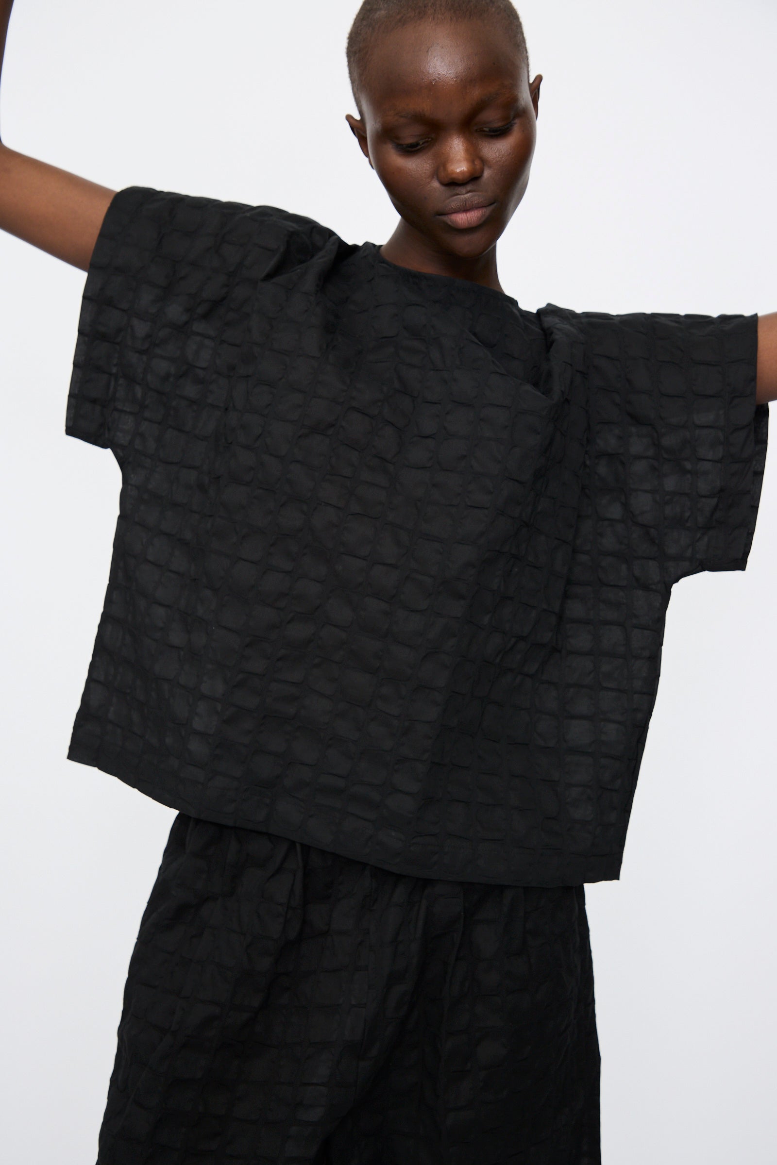A person wears the Black Crane Ripple Finish Top in Black, a loose-fitting, textured Japanese cotton shirt, standing with arms slightly raised against a plain light background.