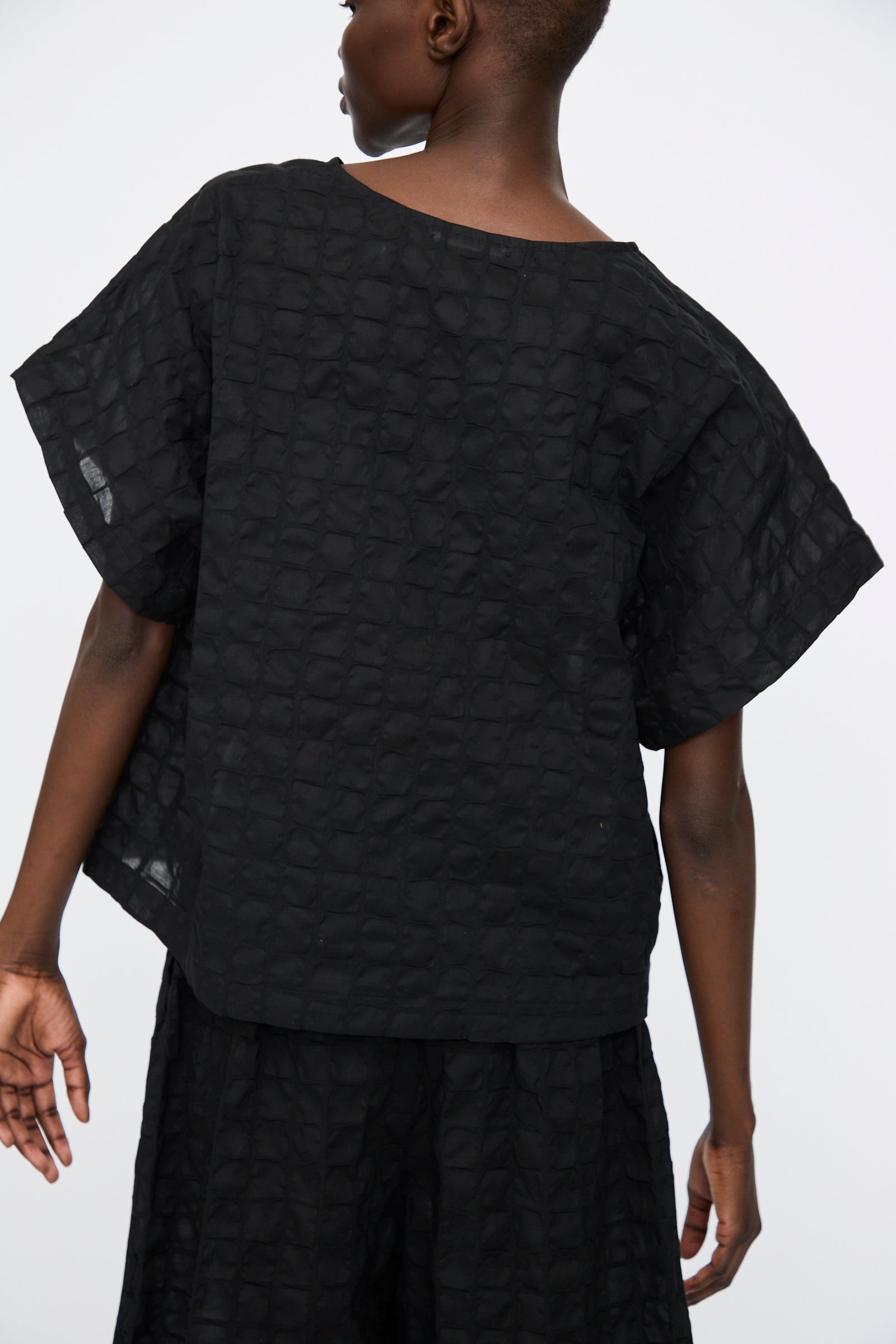 Person wearing the Black Crane Ripple Finish Top in Black— a loose-fitting, short-sleeved boxy shirt made from textured Japanese cotton— paired with matching black bottoms, shown from the back against a plain background.