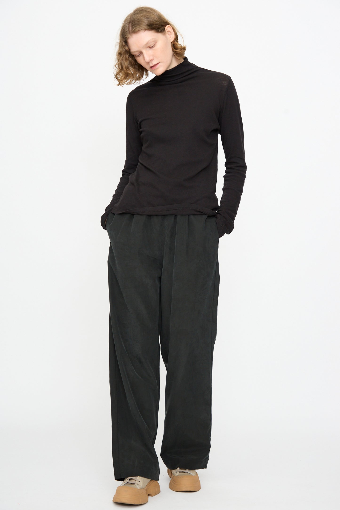 A person stands against a plain white background with hands in pockets, looking down, wearing Black Crane’s Straight Pants in Black, styled with a black turtleneck and tan shoes.
