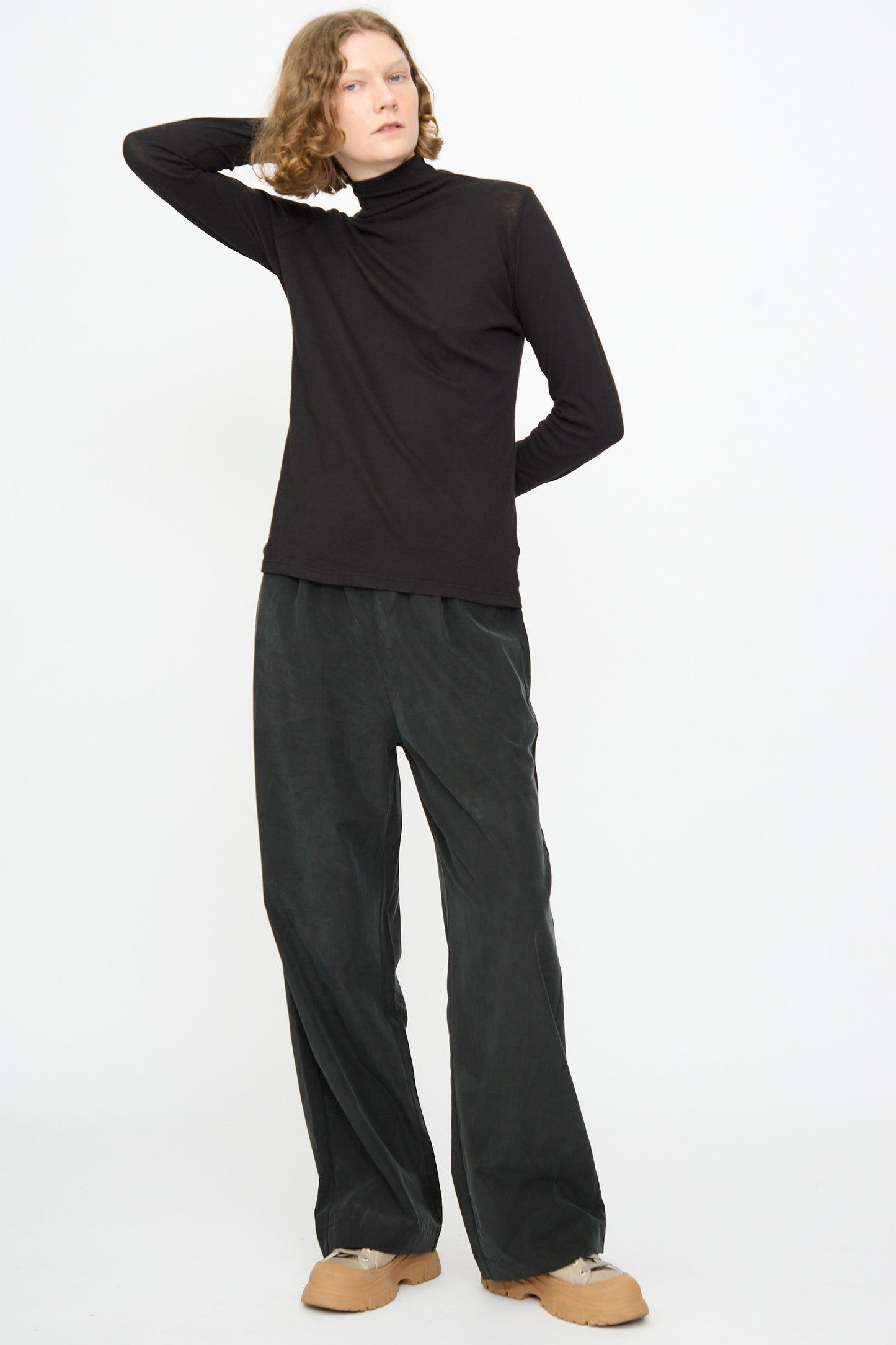 Against a white background, a person stands wearing Black Crane’s Straight Pants in Black—a relaxed-fit style crafted from a soft cotton cupro blend—paired with a black turtleneck and tan shoes. One arm is raised behind their head.