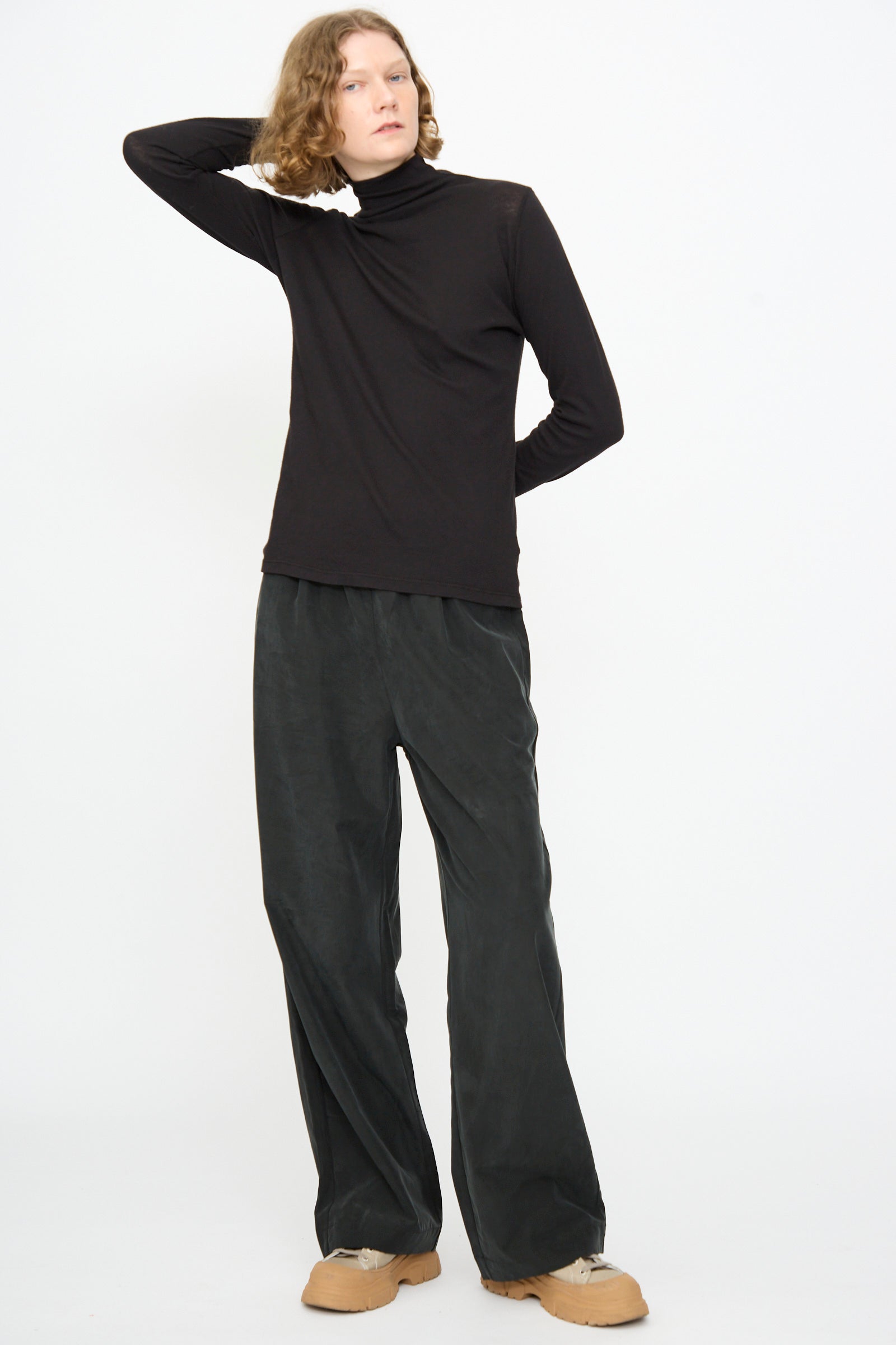 Against a white background, a person stands wearing Black Crane’s Straight Pants in Black—a relaxed-fit style crafted from a soft cotton cupro blend—paired with a black turtleneck and tan shoes. One arm is raised behind their head.