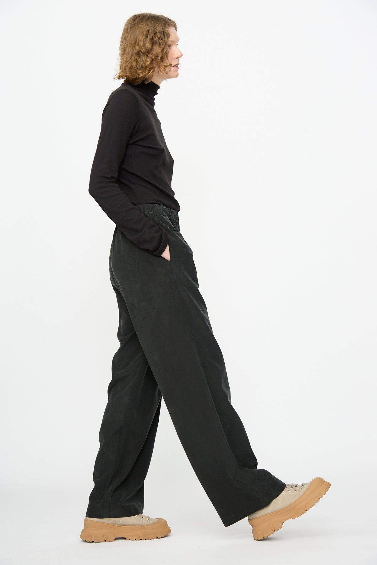 A person stands in profile against a plain white background wearing Black Crane’s Straight Pants in Black, featuring a relaxed fit and crafted from a cotton-cupro blend, paired with a black turtleneck and beige platform shoes.