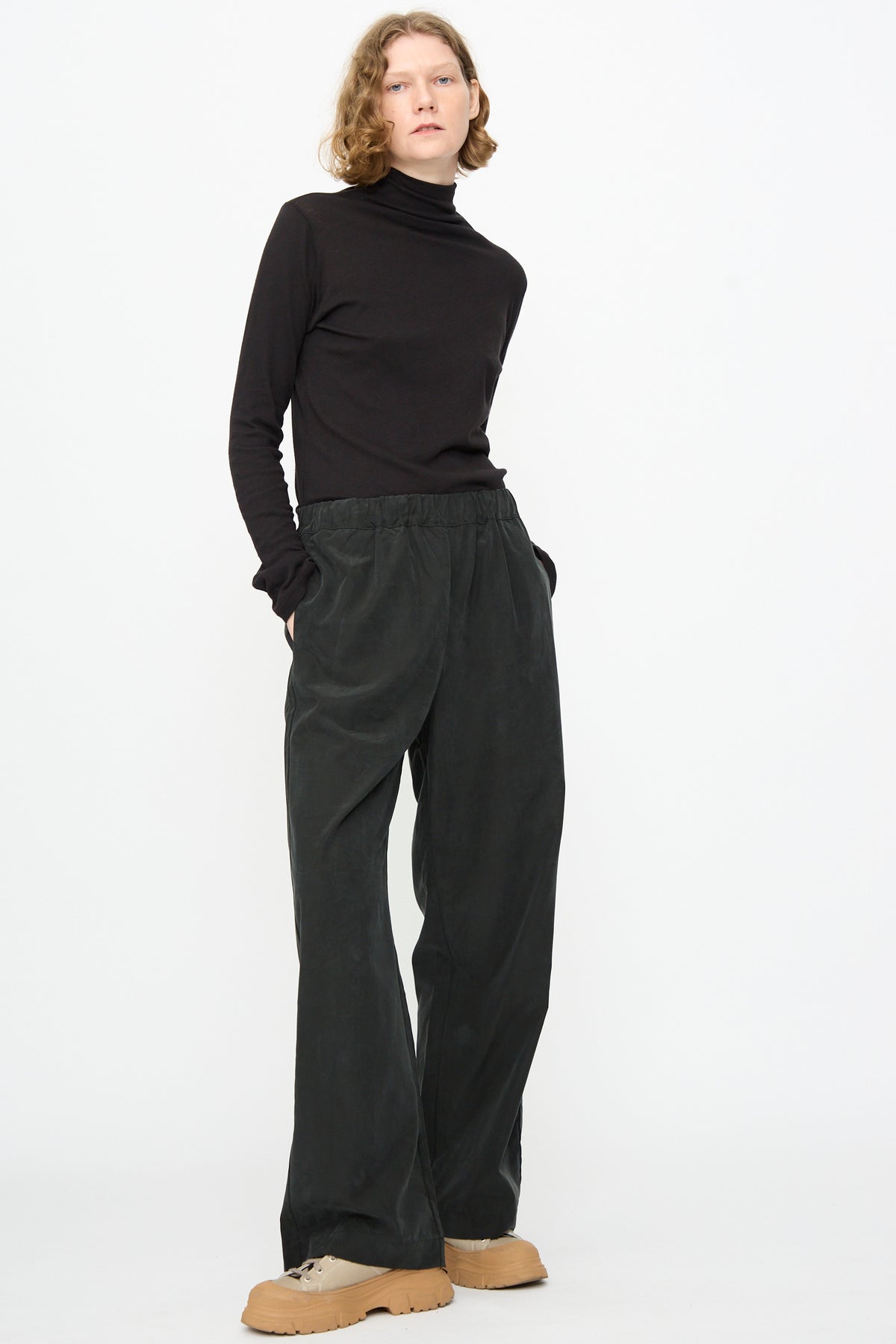 Standing against a white background, the person wears a black turtleneck and Black Crane’s Straight Pants in Black—loose, straight-leg, relaxed fit in a cotton cupro blend—paired with tan platform shoes. Their hands are in their pockets and they have a neutral expression.