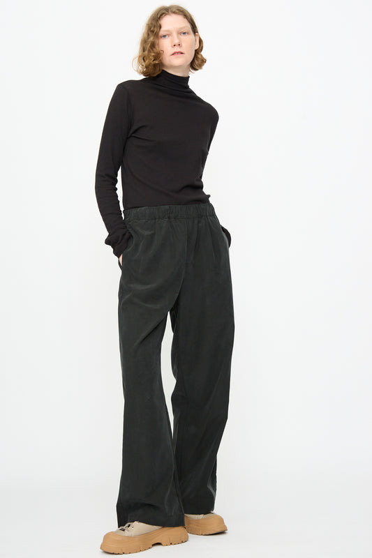 Standing against a white background, the person wears a black turtleneck and Black Crane’s Straight Pants in Black—loose, straight-leg, relaxed fit in a cotton cupro blend—paired with tan platform shoes. Their hands are in their pockets and they have a neutral expression.