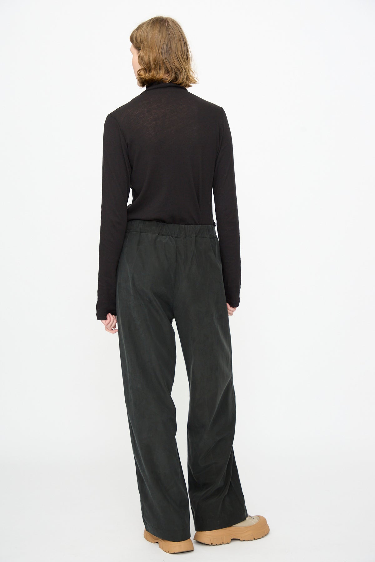 A person stands with their back to the camera, wearing a long-sleeve black top, Black Crane’s Straight Pants in Black—relaxed fit and crafted from a cotton-cupro blend—and tan shoes, set against a plain white background.