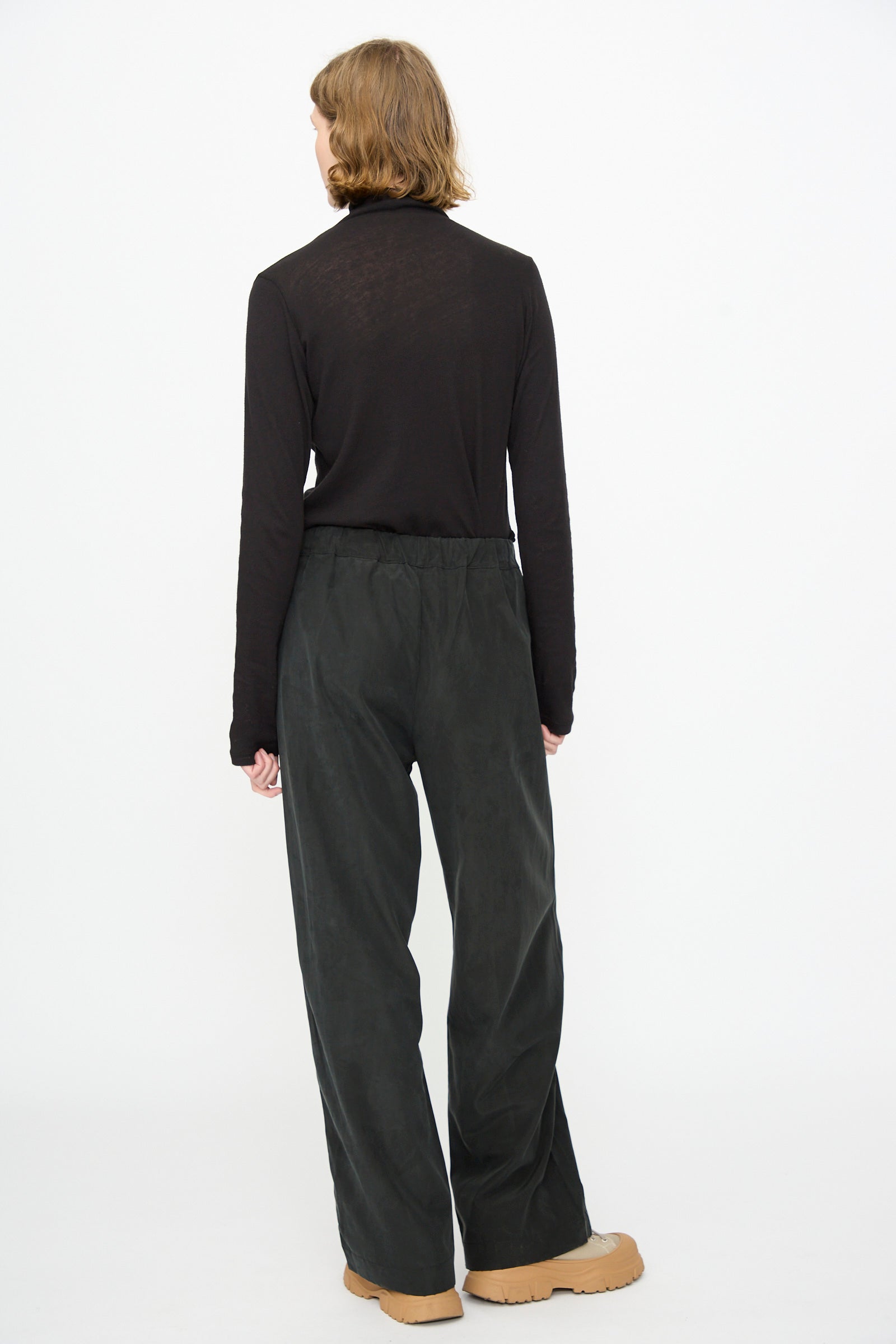 A person stands with their back to the camera, wearing a long-sleeve black top, Black Crane’s Straight Pants in Black—relaxed fit and crafted from a cotton-cupro blend—and tan shoes, set against a plain white background.