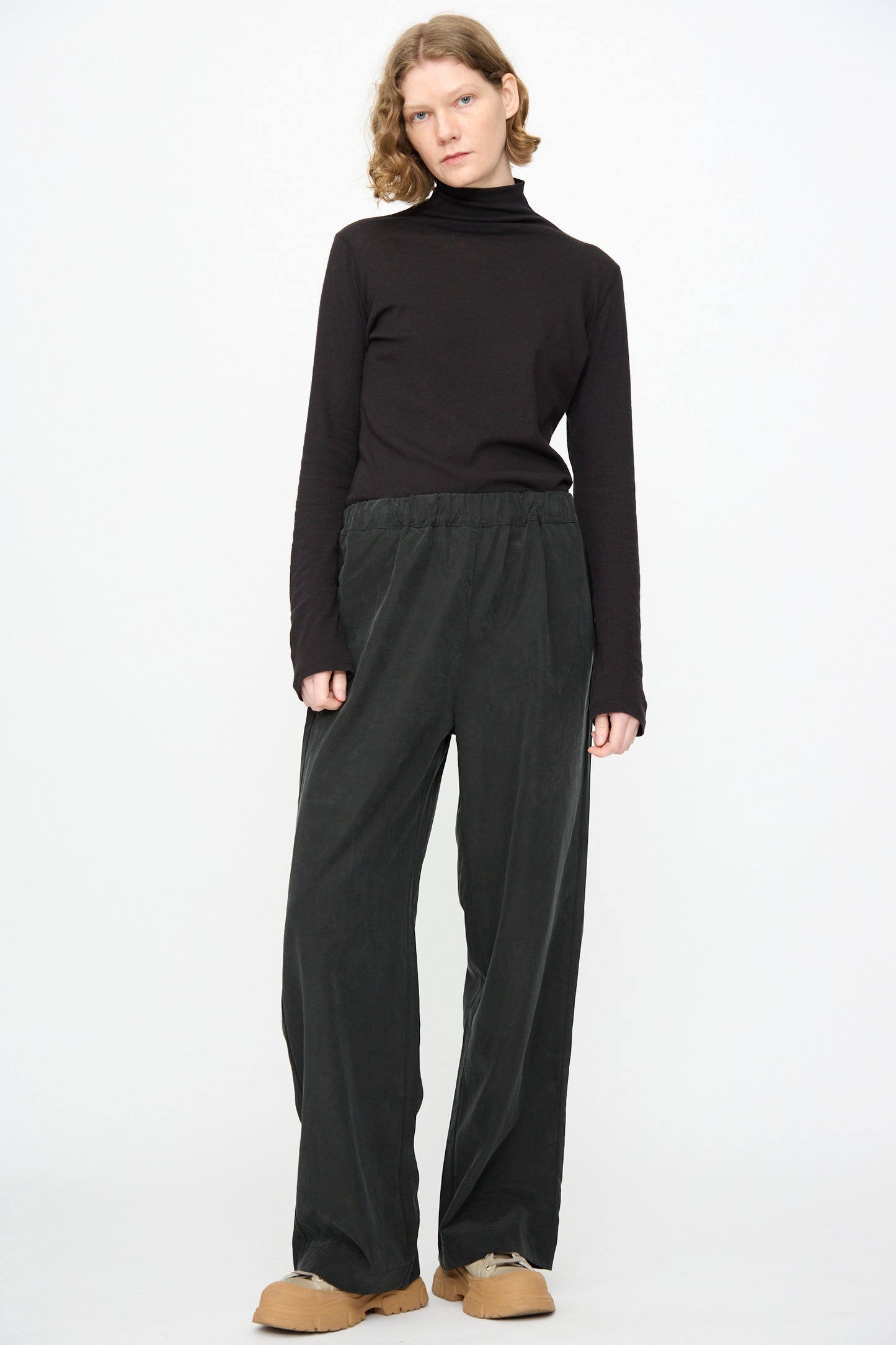 A person stands against a white background wearing a black turtleneck, Black Crane Straight Pants in Black made from a cotton cupro blend with a relaxed fit, and tan shoes.