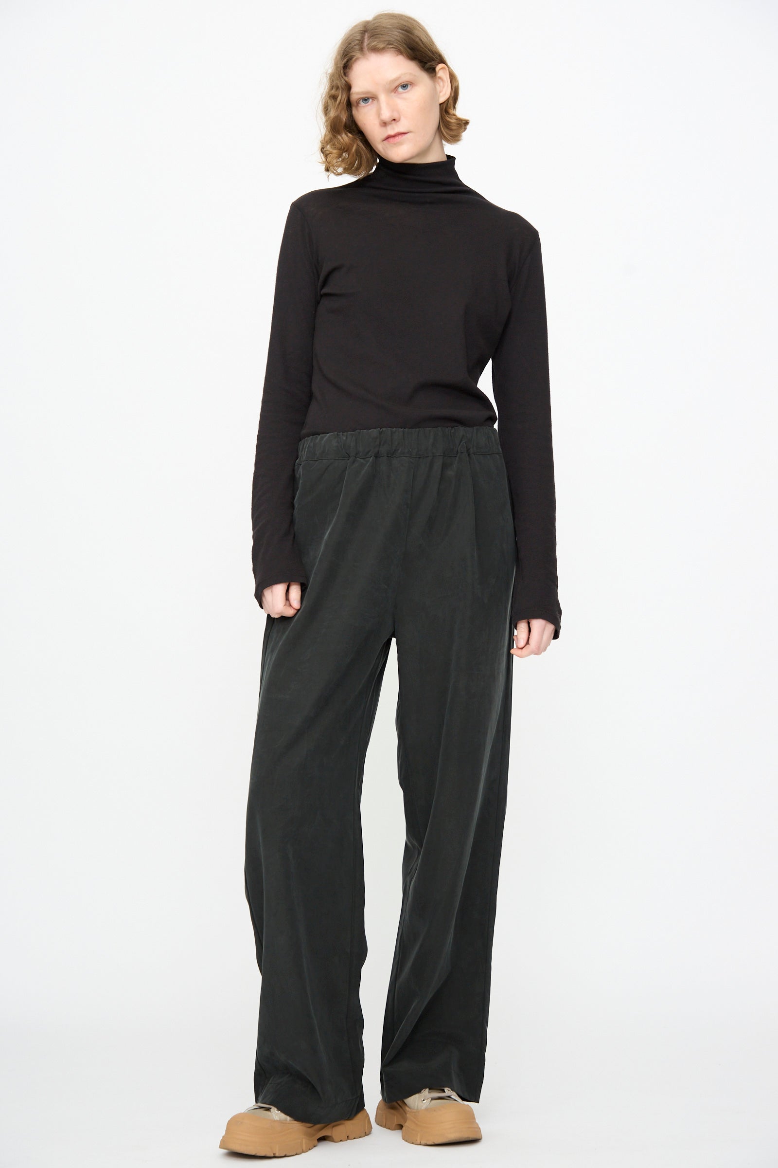 A person stands against a white background wearing a black turtleneck, Black Crane Straight Pants in Black made from a cotton cupro blend with a relaxed fit, and tan shoes.