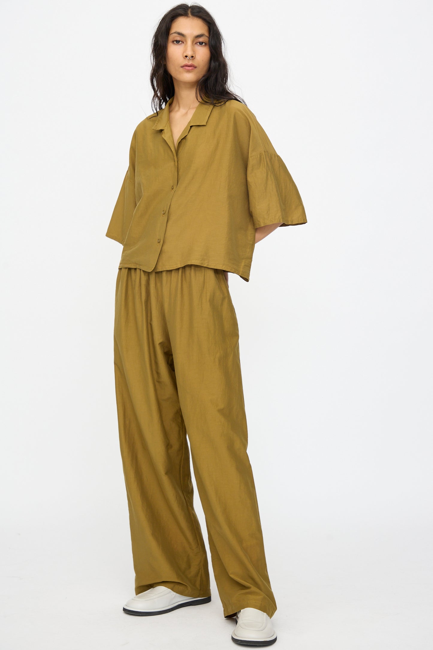 A person wears Black Crane's Straight Pants in Dark Olive, styled with a loose olive-brown button-up shirt and white slip-on shoes, standing against a plain white background.