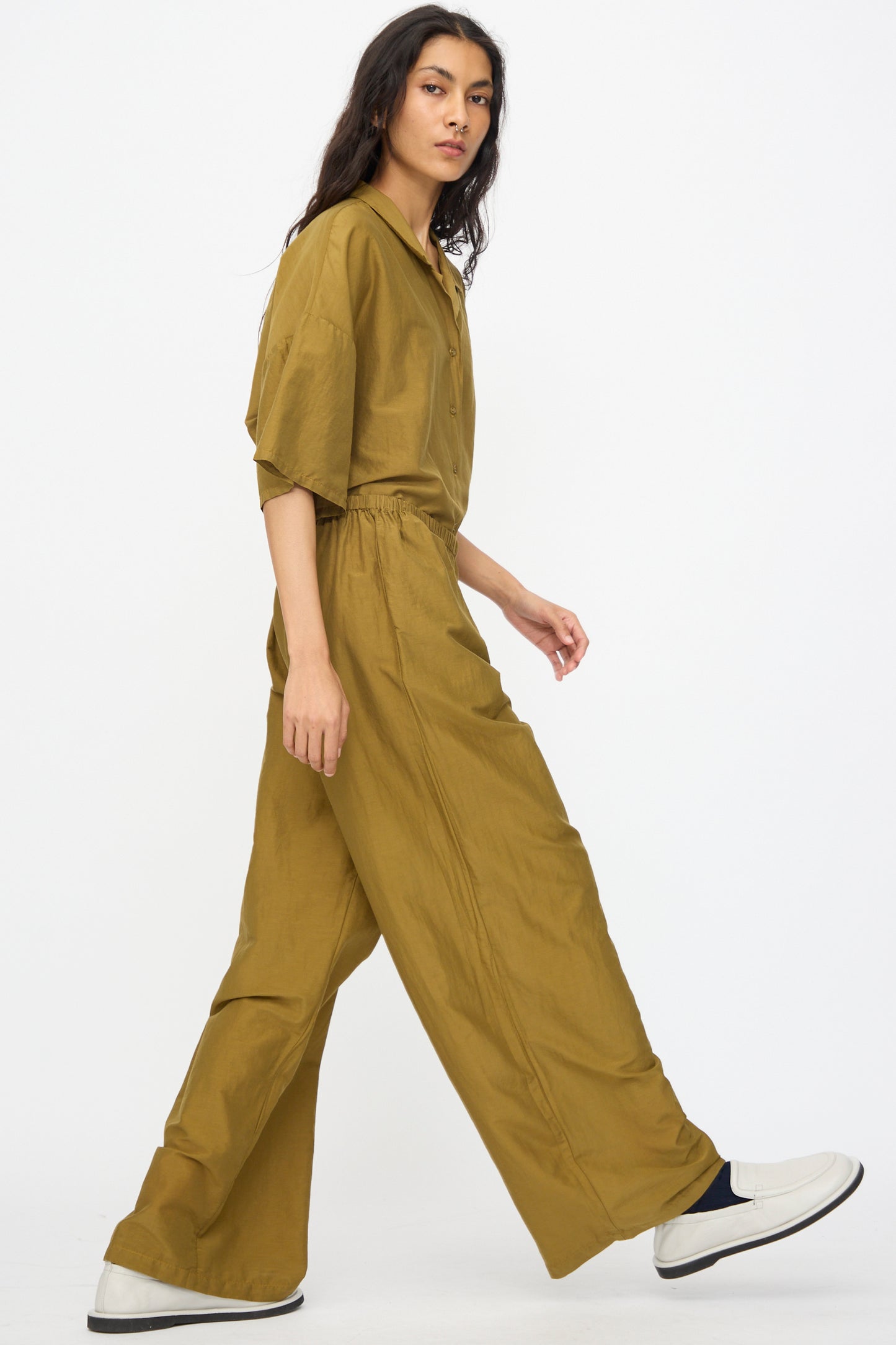 A person with long dark hair walks against a plain white background, wearing Black Crane’s Straight Pants in Dark Olive, a loose-fitting olive-green shirt, and white slip-on shoes.