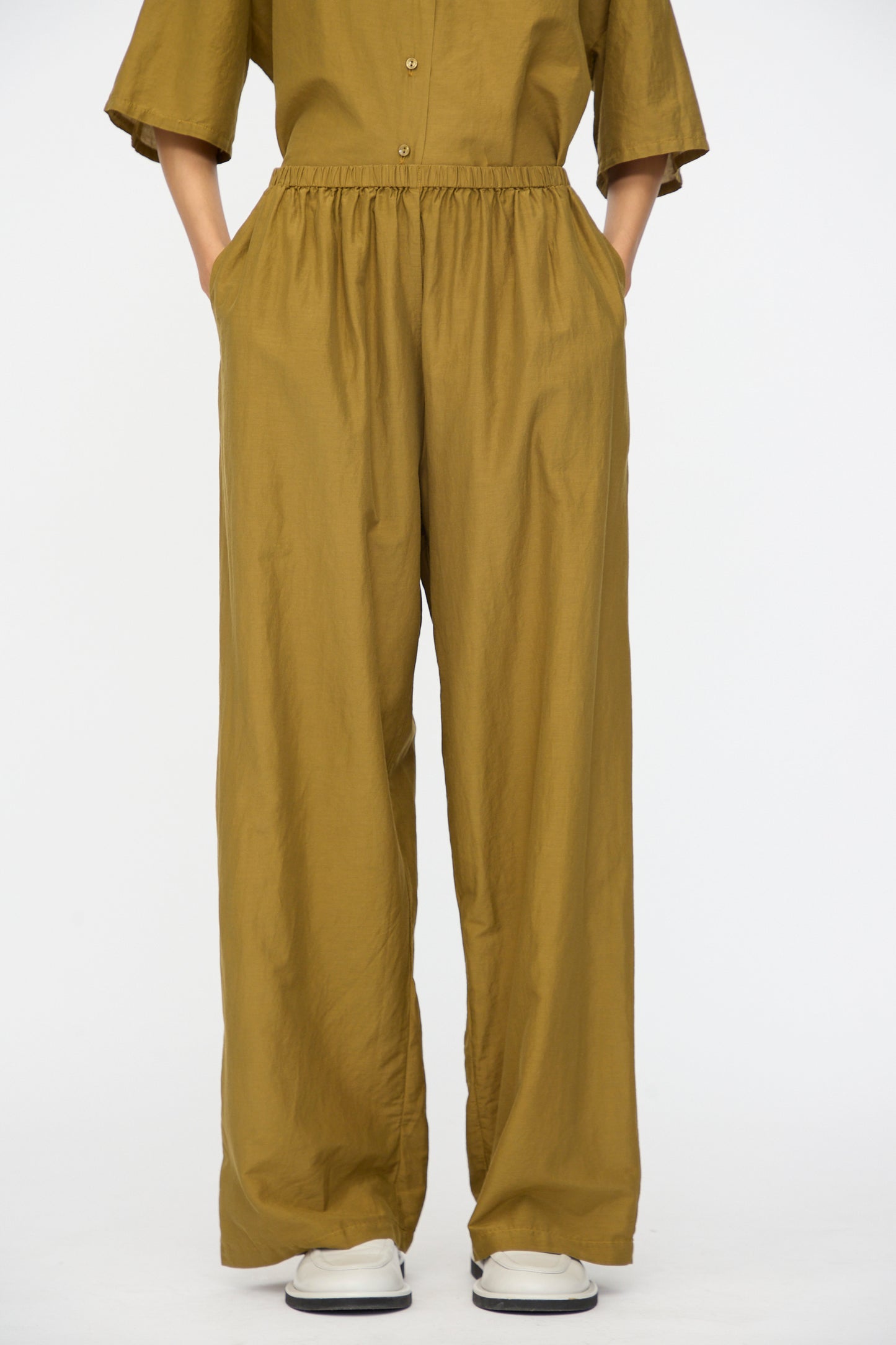 Person wearing Black Crane Straight Pants in Dark Olive with hands in pockets, white shoes, and a matching buttoned top, standing against a plain white background.