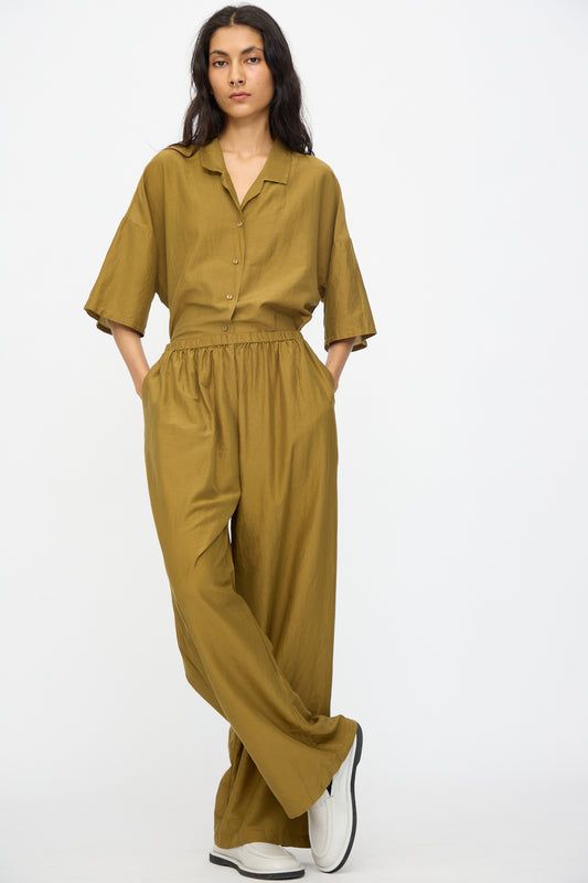 A person stands with hands in pockets, wearing Black Crane Straight Pants in Dark Olive and a relaxed fit olive green button-up shirt, paired with white shoes against a plain white background.