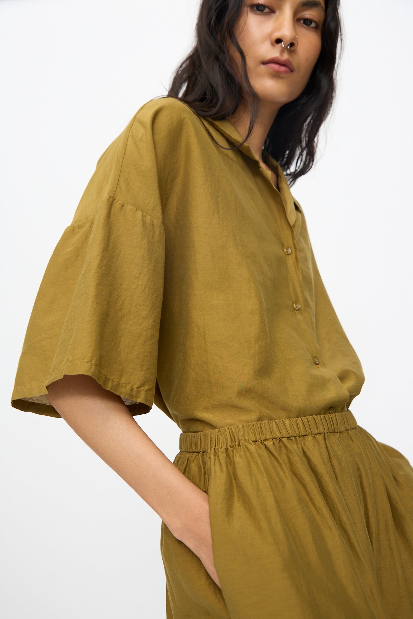 A woman wearing Black Crane’s Straight Pants in Dark Olive with a matching loose olive-green button-up shirt stands against a plain background, one hand in her pocket.