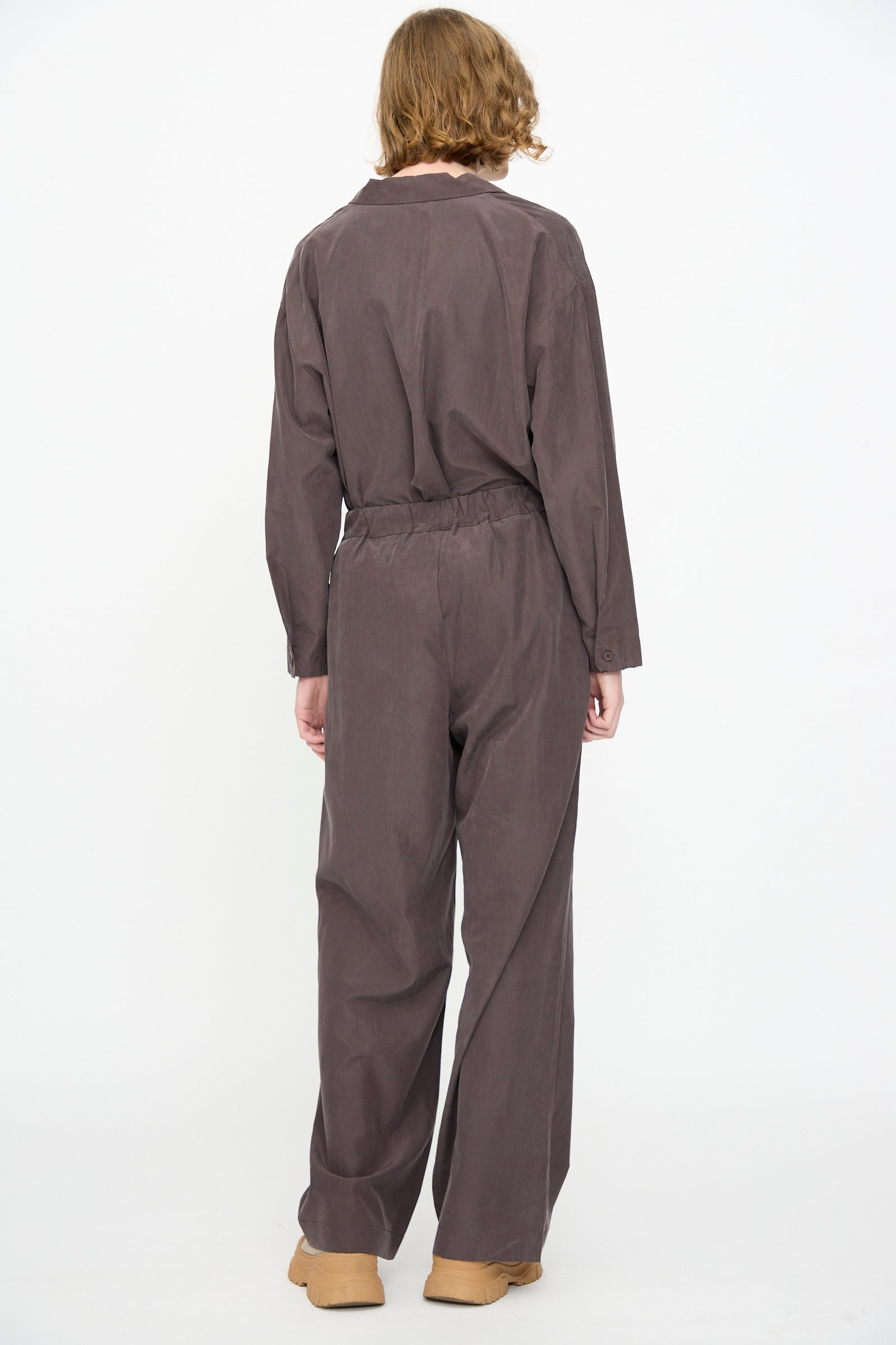 A person is shown from the back, standing against a white background and wearing Black Crane’s loose-fitting Straight Pants in Dark Plum, made from a cotton-cupro blend with a relaxed fit and straight leg, paired with tan shoes.