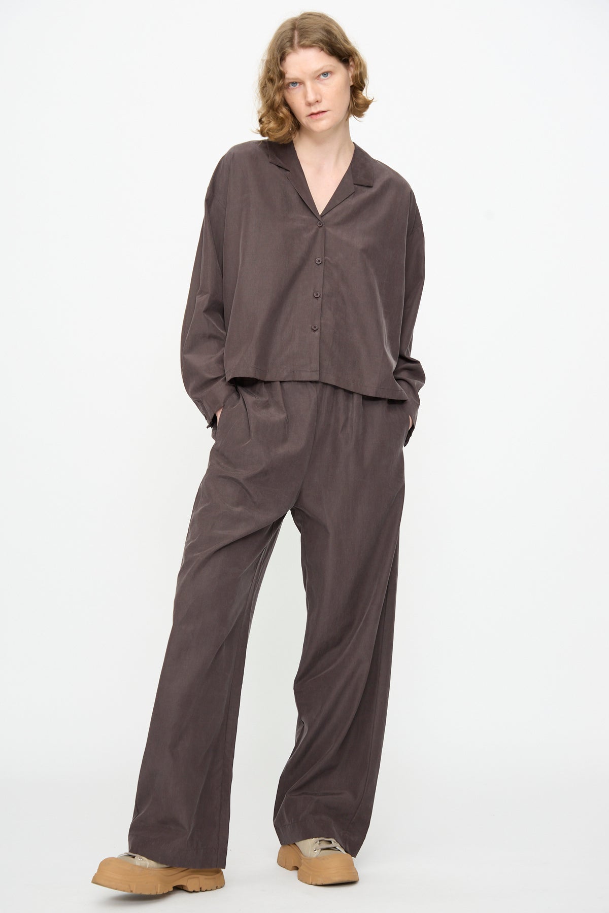 A person wearing a loose-fitting, dark brown button-up shirt crafted from a comfortable cotton cupro blend and Black Crane’s Straight Pants in Dark Plum, paired with light brown shoes, stands against a plain white background.