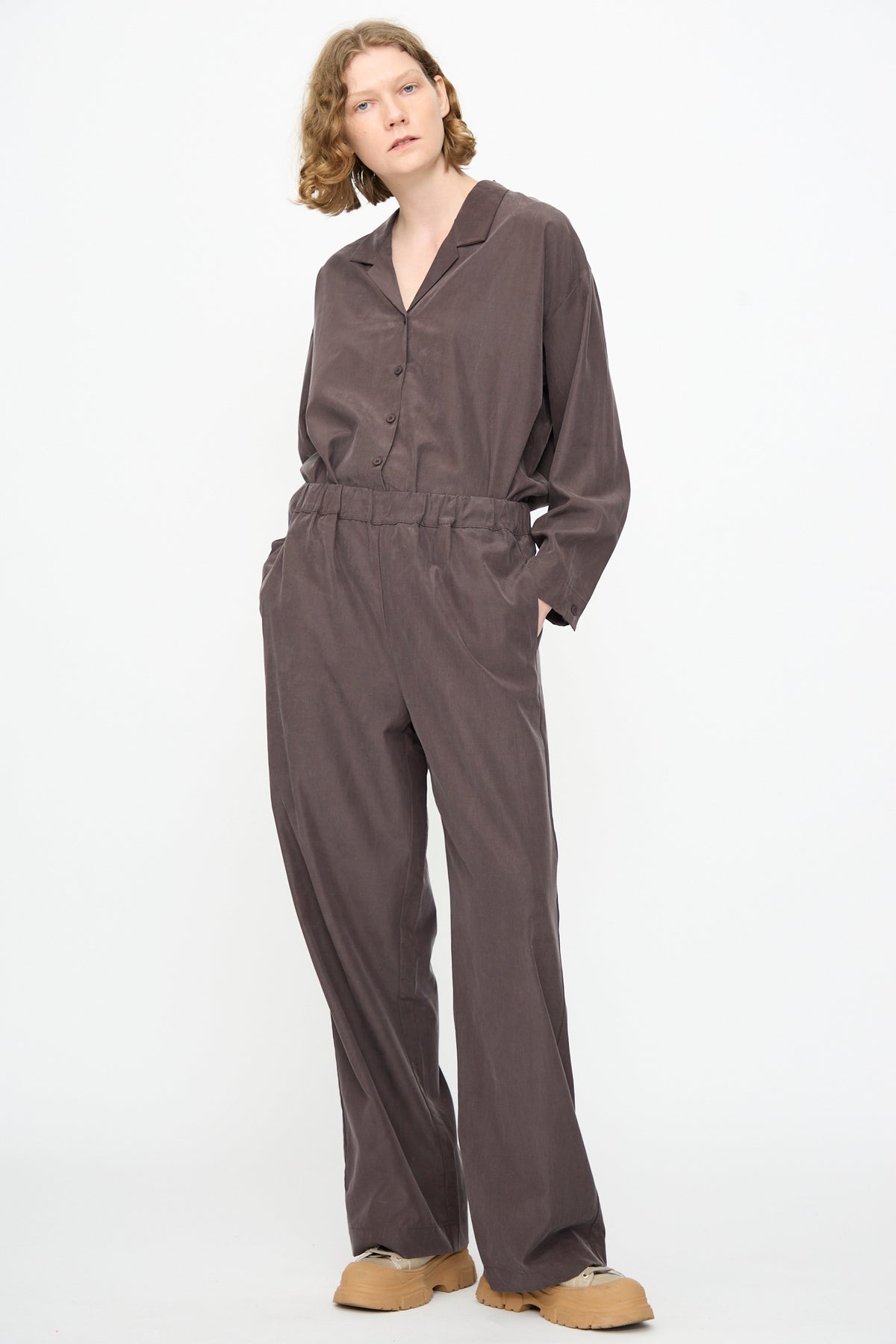 A person stands against a white background wearing Black Crane’s relaxed fit Straight Pants in Dark Plum and a loose-fitting brown button-up shirt crafted from a soft cotton cupro blend, with hands in pockets and tan shoes.