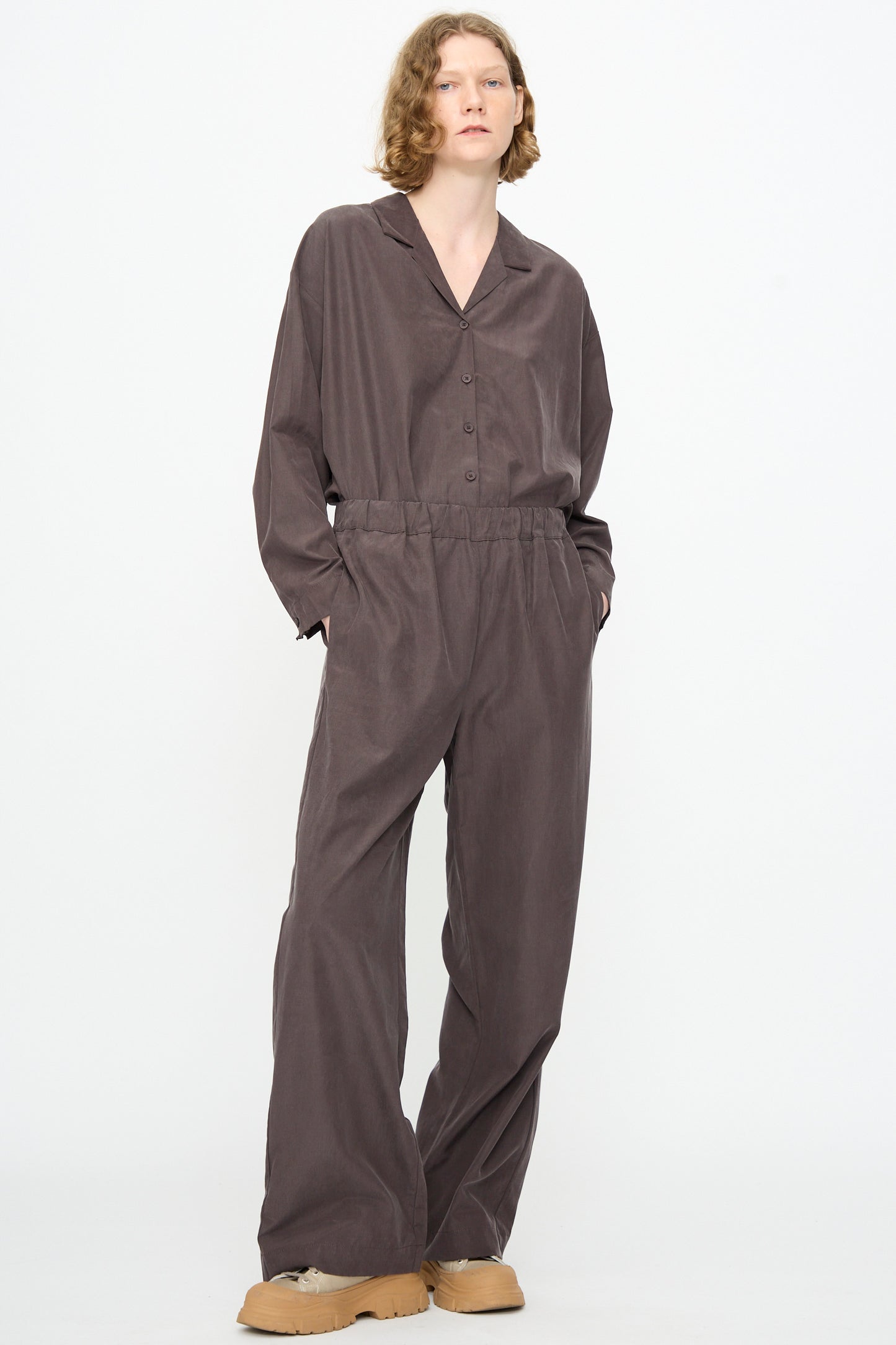 A person with short, wavy hair wears the Black Crane Straight Pants in Dark Plum, crafted from a cotton cupro blend and designed with a relaxed fit and straight legs. Paired effortlessly with tan platform shoes, they stand against a plain white background with their hands in their pockets.