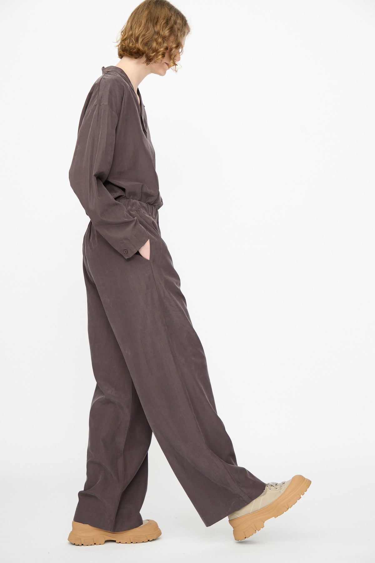 A person wearing Black Crane’s Straight Pants in Dark Plum, made from a soft cotton cupro blend, stands sideways with one foot slightly forward against a plain white background, paired with tan platform shoes.