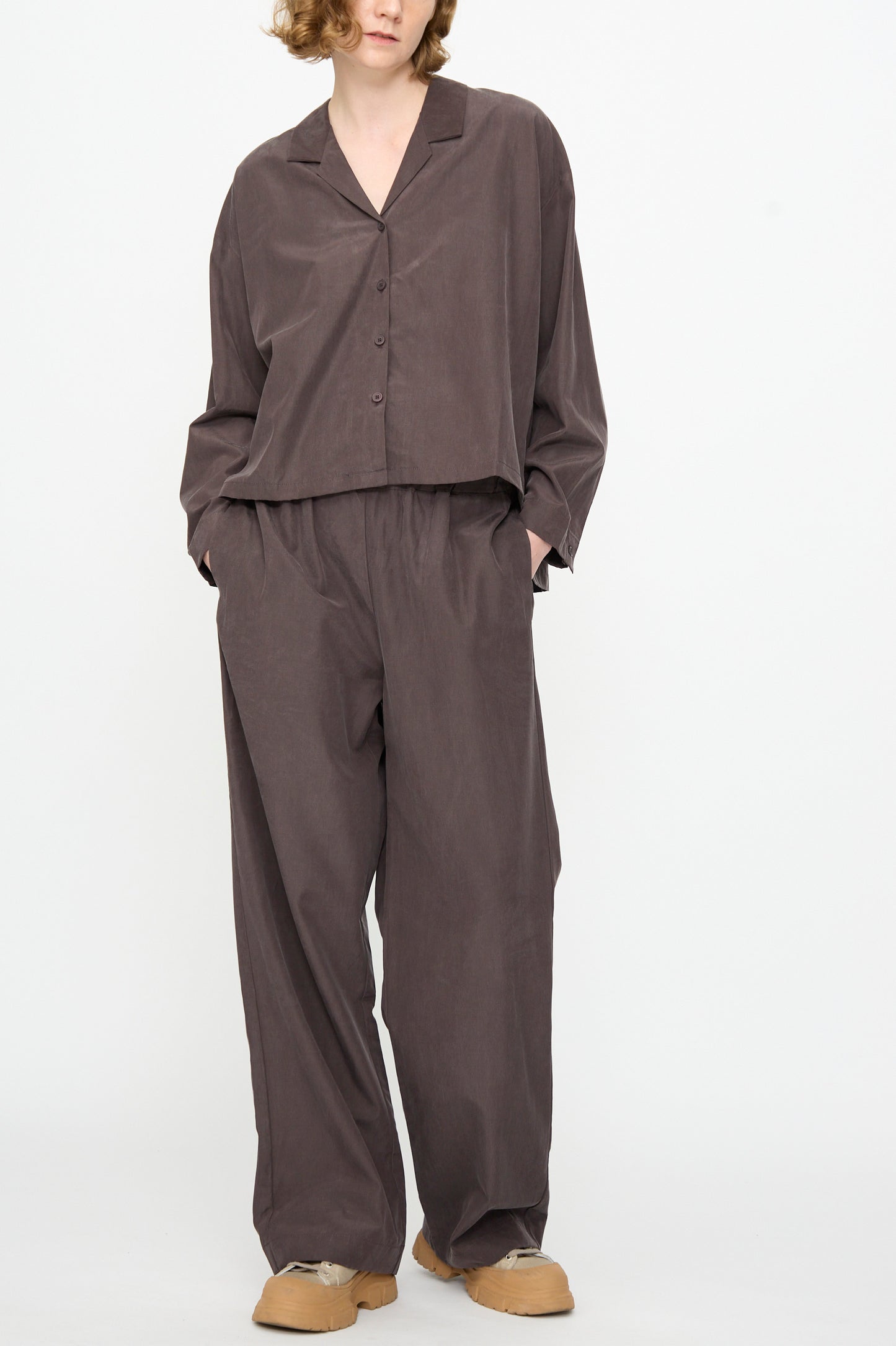 A person wearing Black Crane’s Straight Pants in Dark Plum—a relaxed fit, straight pant in a soft cotton cupro blend—with hands in pockets, standing against a plain white background.