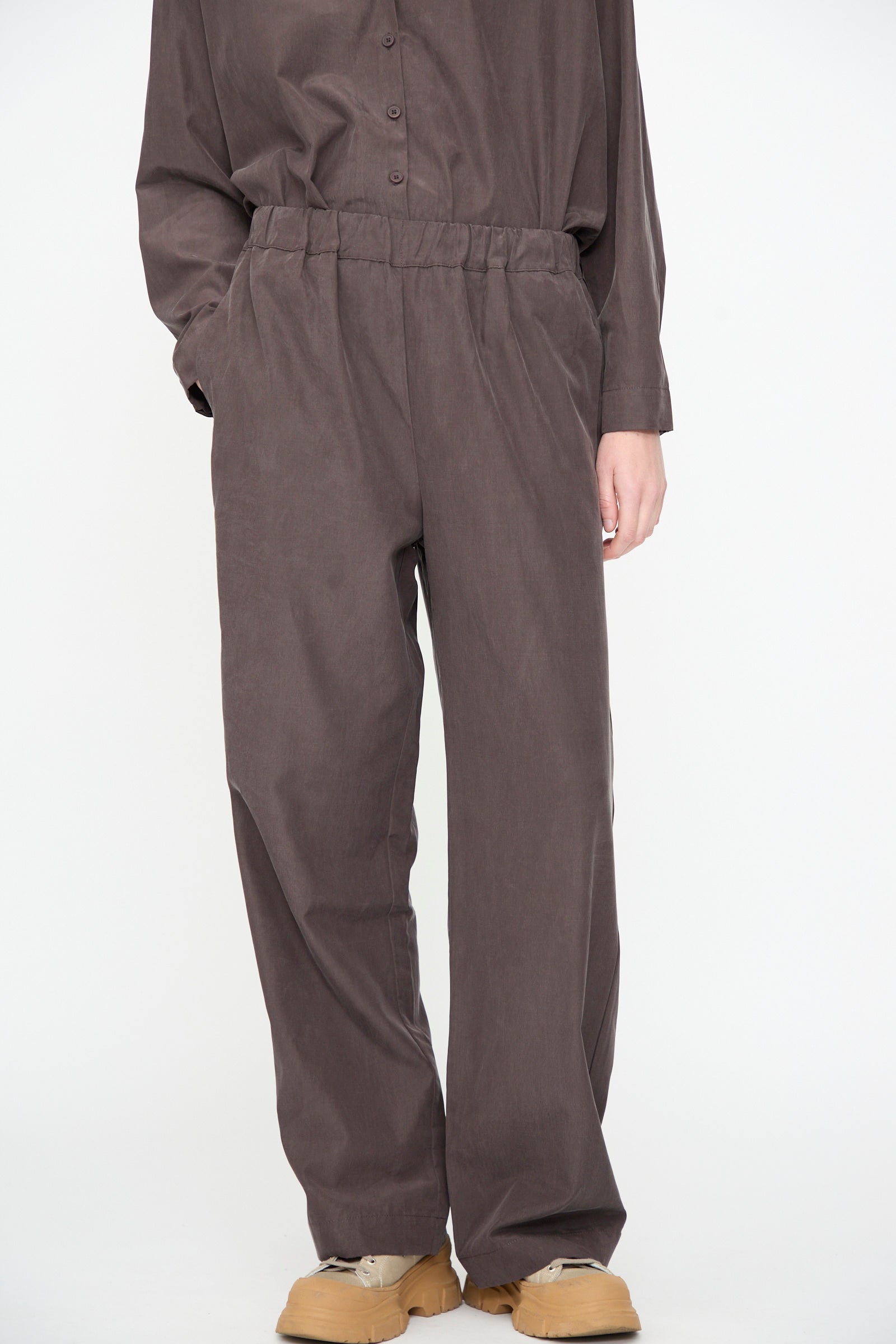 Person wearing loose, relaxed fit Straight Pants in Dark Plum by Black Crane and a matching button-up shirt, with hands in pockets, standing against a plain white background. Only the lower body is visible. The straight pant is crafted from a comfortable cotton cupro blend.