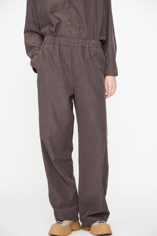Person wearing loose, relaxed fit Straight Pants in Dark Plum by Black Crane and a matching button-up shirt, with hands in pockets, standing against a plain white background. Only the lower body is visible. The straight pant is crafted from a comfortable cotton cupro blend.