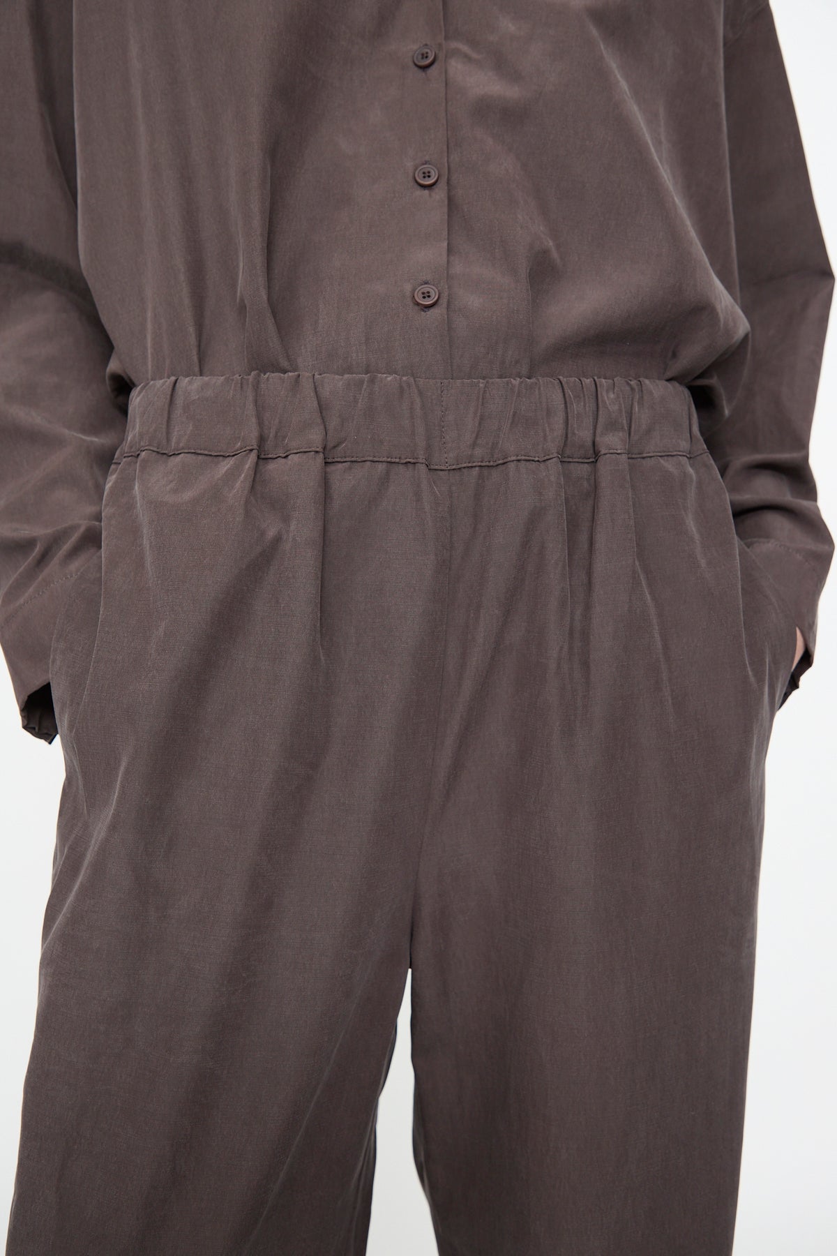 A person wears the Black Crane Straight Pants in Dark Plum, featuring a relaxed fit and straight leg, crafted from a soft cotton cupro blend. Their hands are in their pockets, and the photo is cropped at the shoulders and knees.