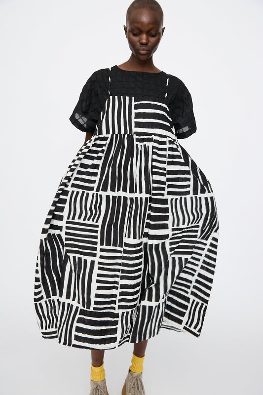 A person wears the Black Crane Strap Dress in Black Print—a loose, graphic midi dress with a solid black textured top—paired with yellow socks and beige Japanese cotton shoes, standing against a plain white background.