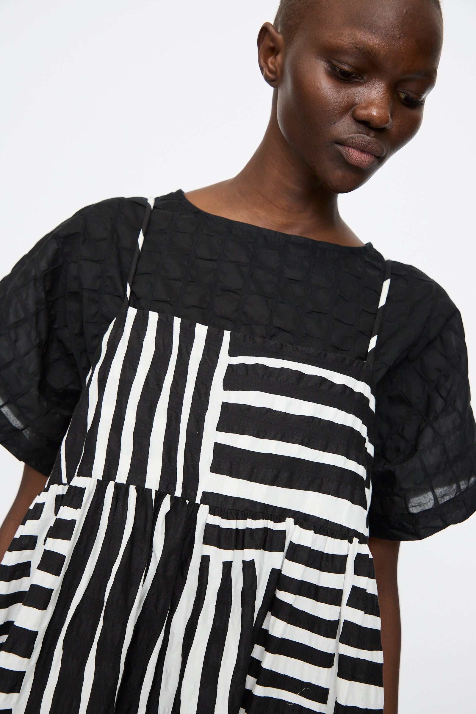 A person wears the Black Crane Strap Dress in Black Print, a sleeveless midi with black and white graphic stripes, layered over a black textured short-sleeve top.