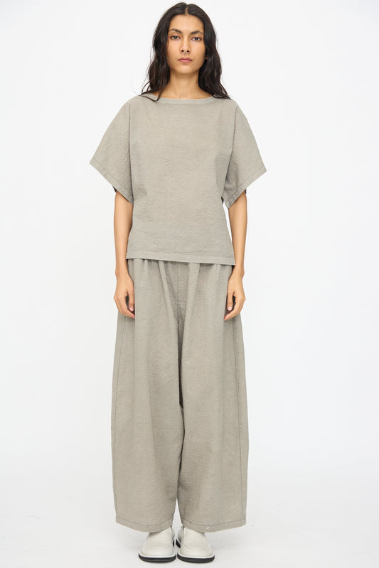 A person stands against a white background wearing the Black Crane Textured Cotton Boxed Top in Pale Grey with wide-leg pants and white shoes.