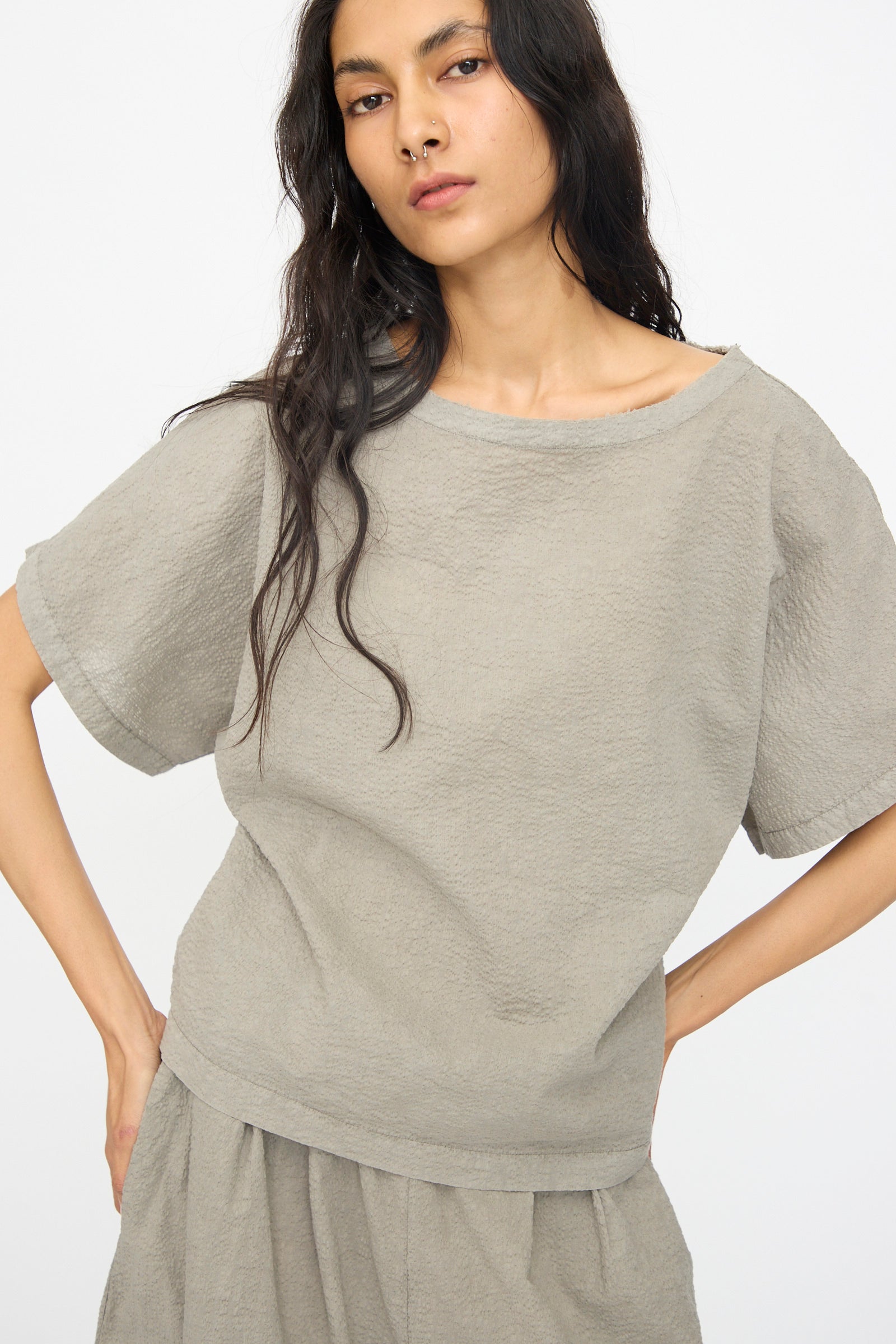 A woman with long dark hair models Black Crane’s Textured Cotton Boxed Top in Pale Grey, paired with a matching skirt, against a plain white background.