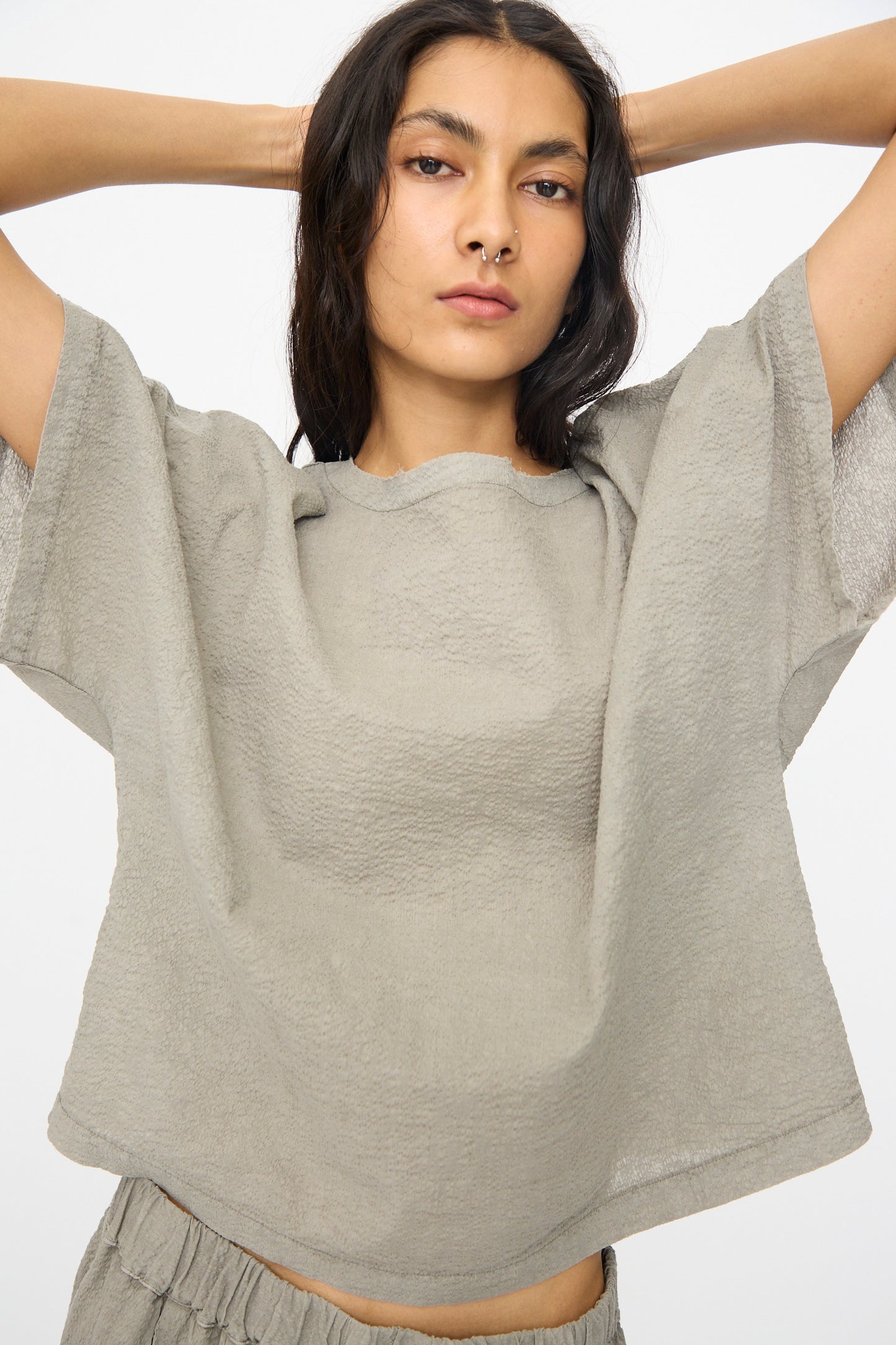 A woman in the Black Crane Textured Cotton Boxed Top in Pale Grey, styled with matching bottoms and a neutral expression, stands with raised arms against a plain white background.