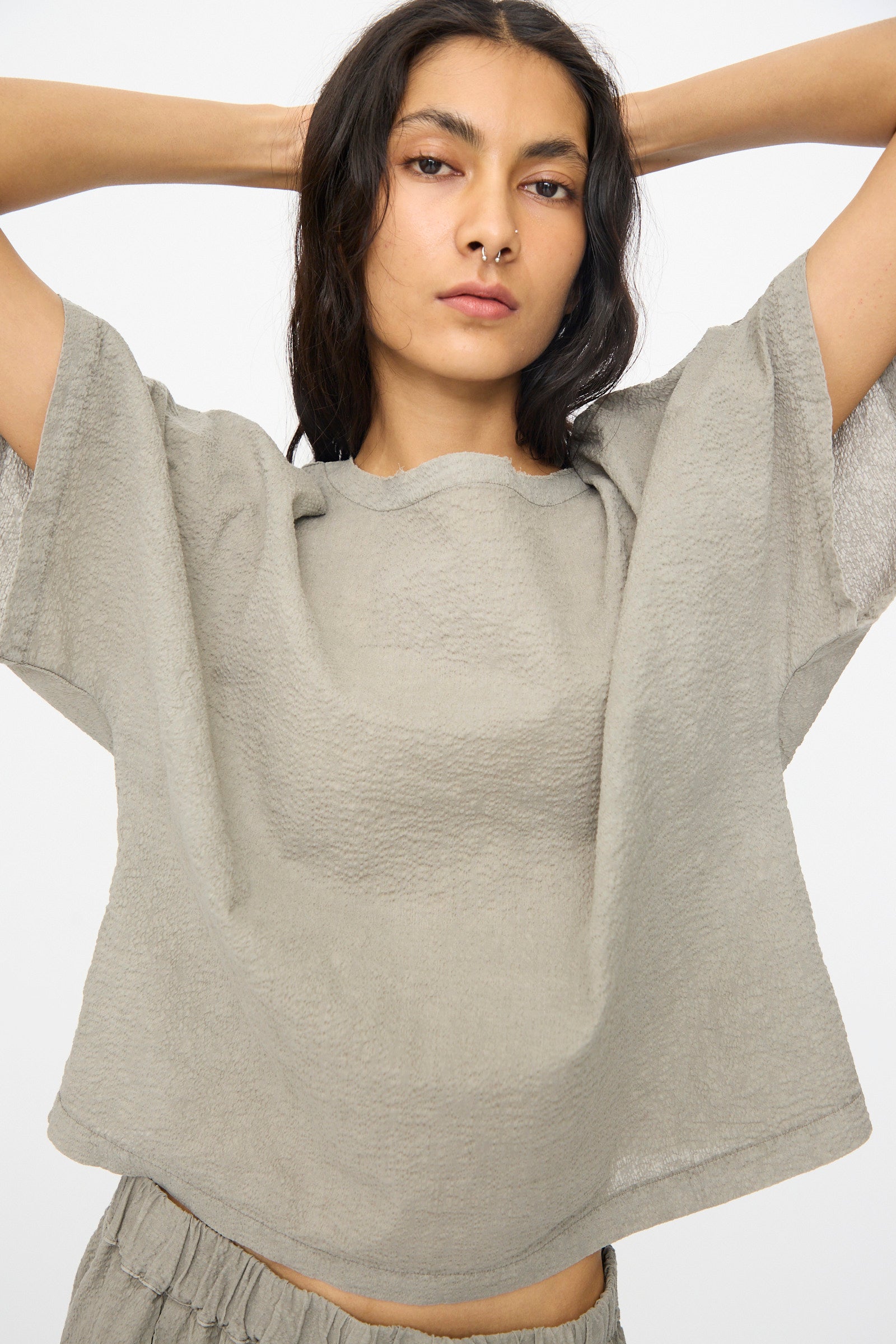 A woman in the Black Crane Textured Cotton Boxed Top in Pale Grey, styled with matching bottoms and a neutral expression, stands with raised arms against a plain white background.