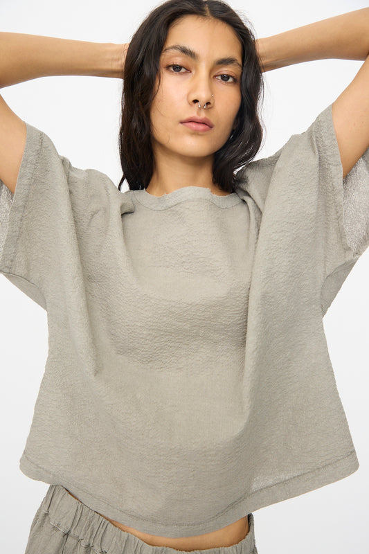 A woman in the Black Crane Textured Cotton Boxed Top in Pale Grey, styled with matching bottoms and a neutral expression, stands with raised arms against a plain white background.