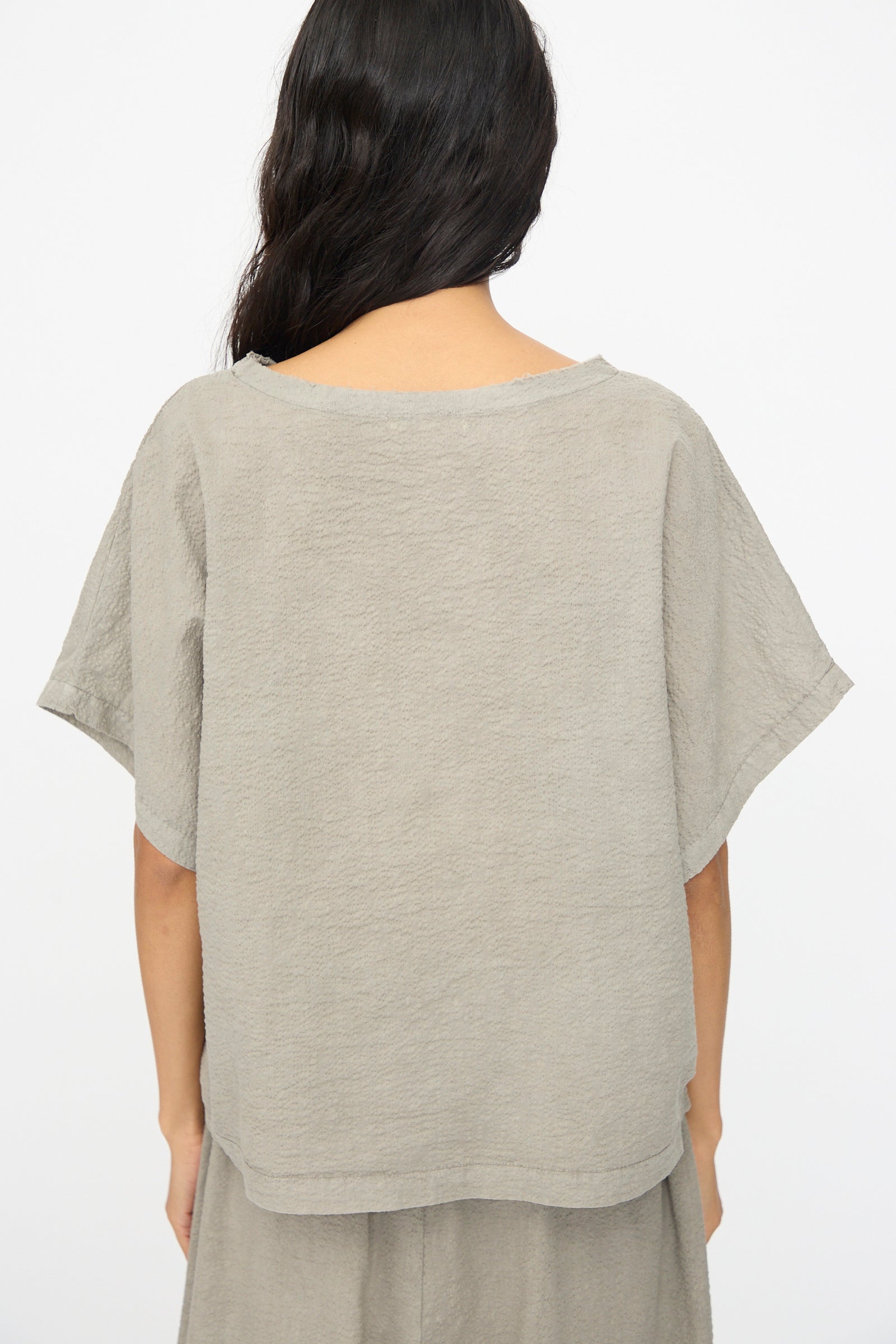 A person with long dark hair is shown from the back wearing the Black Crane Textured Cotton Boxed Top in Pale Grey, featuring short sleeves and Japanese textured cotton, against a plain background.