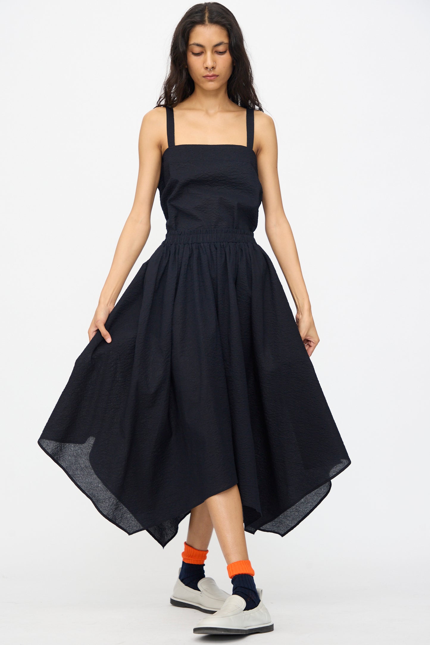 A woman stands against a plain white background wearing the Black Crane Textured Cotton Paper Skirt in Midnight, paired with orange socks and white slip-on shoes.