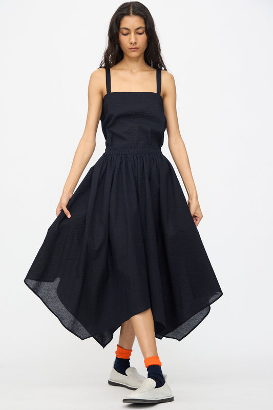 A woman stands against a plain white background wearing the Black Crane Textured Cotton Paper Skirt in Midnight, paired with orange socks and white slip-on shoes.