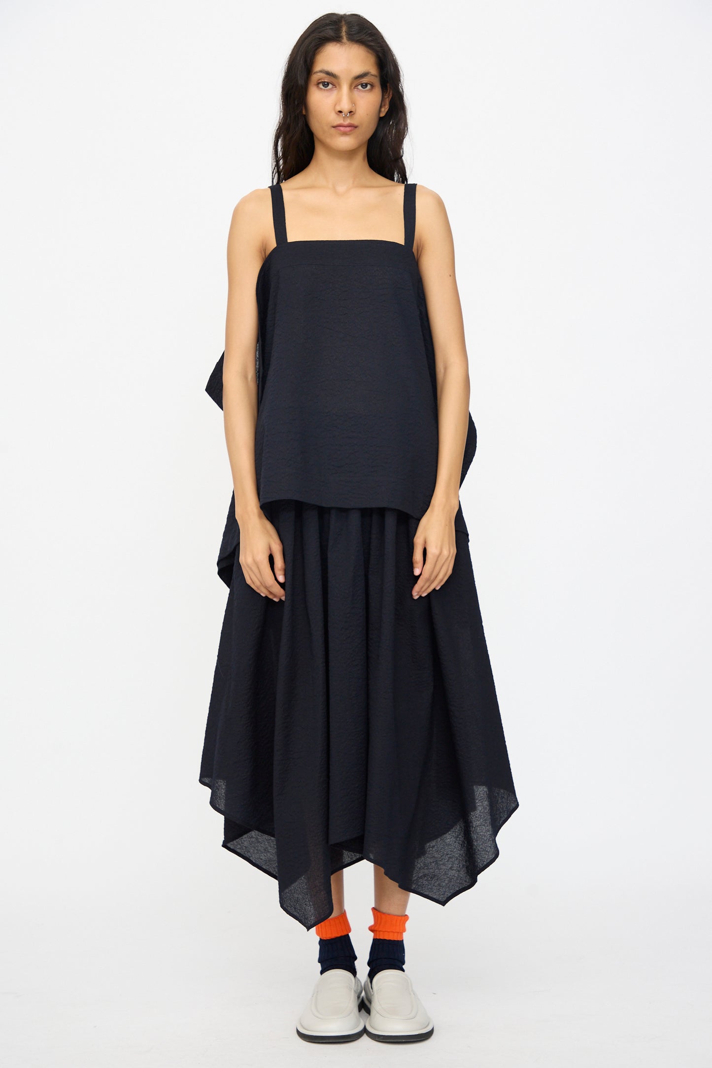 A woman faces forward in a loose black sleeveless top and the Black Crane Textured Cotton Paper Skirt in Midnight, featuring an elastic waistband, paired with orange socks and white slip-on shoes against a plain white background.
