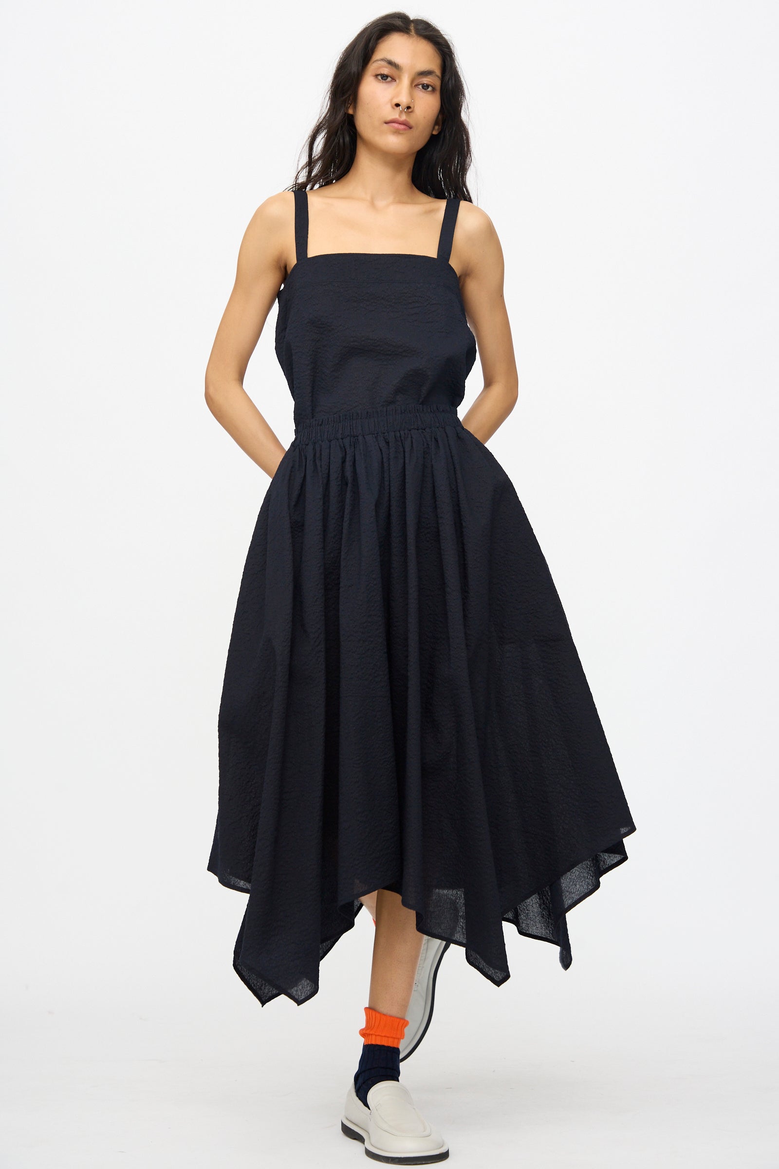 A person wearing the Black Crane Textured Cotton Paper Skirt in Midnight, orange socks, black ankle socks, and white slip-on shoes stands against a plain white background.