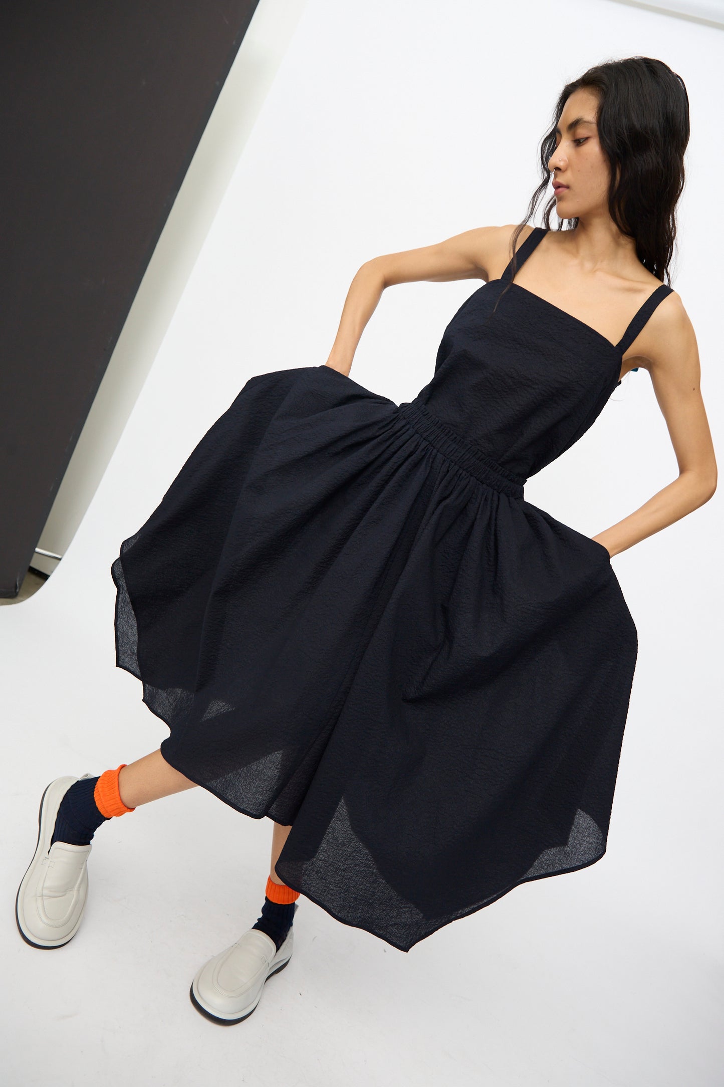 A woman poses in the Black Crane Textured Cotton Paper Skirt in Midnight, featuring an elastic waistband and pockets, paired with white shoes, orange socks, and dark socks, against a plain background.
