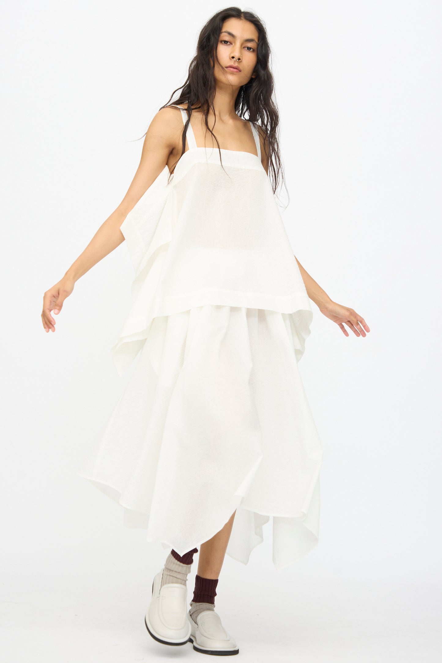 A person wears the Black Crane Textured Cotton Paper Skirt in White, styled loosely with layers, paired with white shoes and maroon socks, standing against a plain white background.