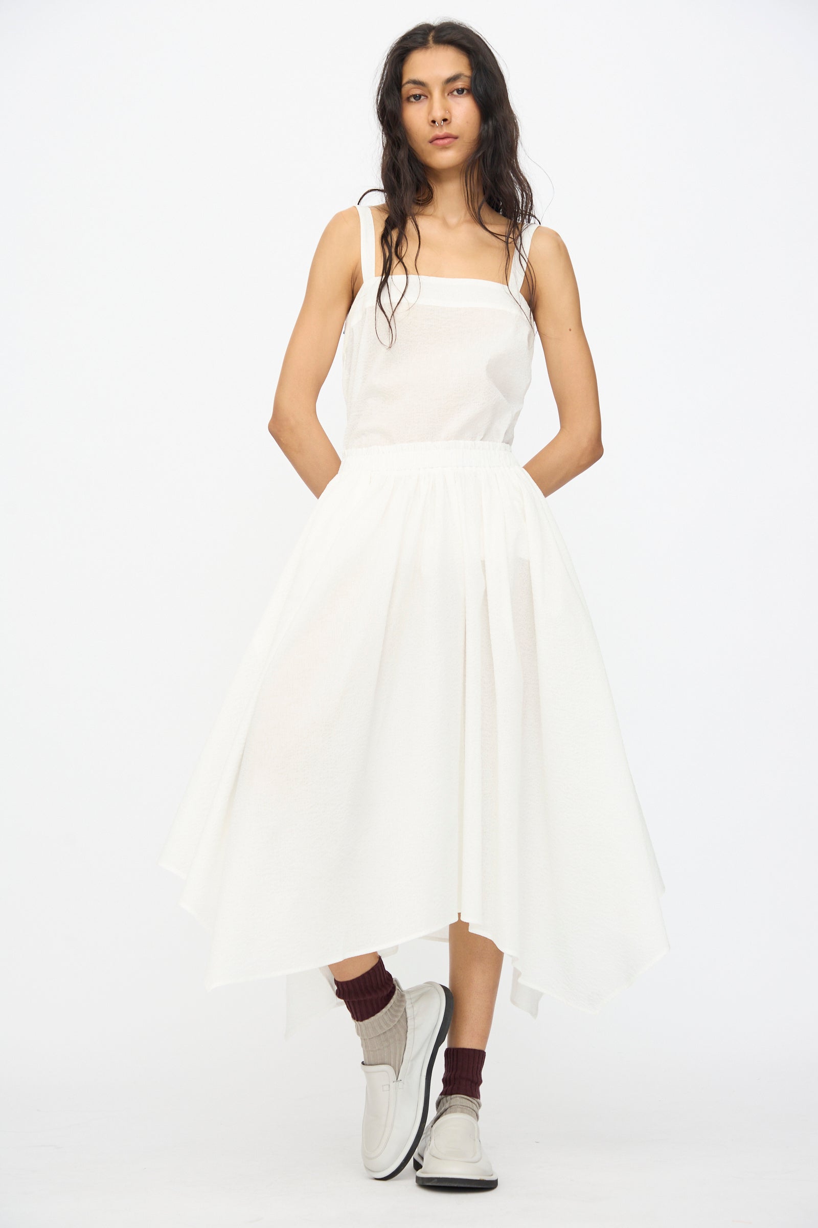 A woman wears the Black Crane Textured Cotton Paper Skirt in White, styled with brown socks and white loafers, standing against a plain white background.