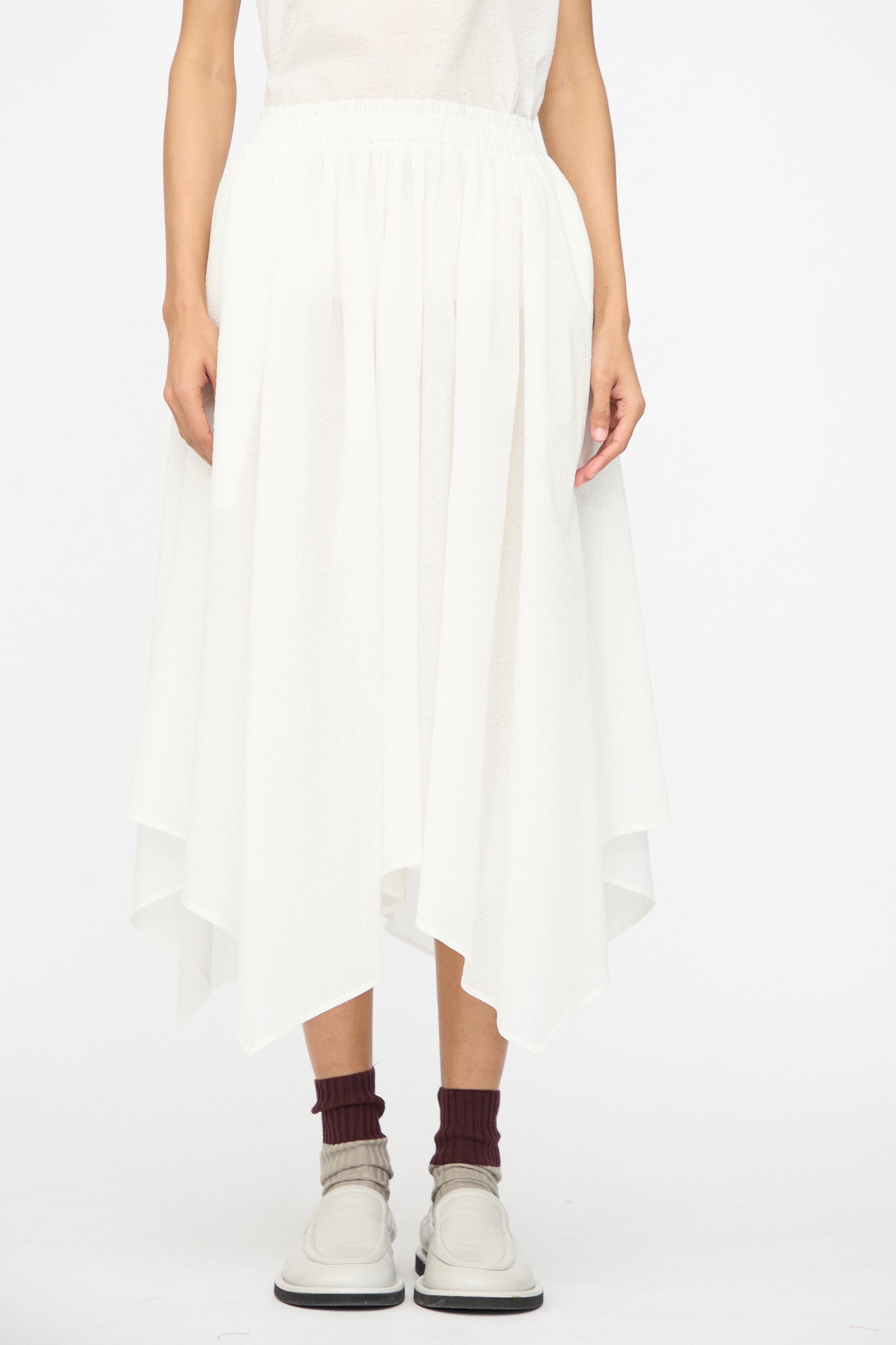 A person wears the Black Crane Textured Cotton Paper Skirt in White, featuring an elastic waistband and Japanese textured cotton, styled with maroon socks and off-white shoes against a plain white background.